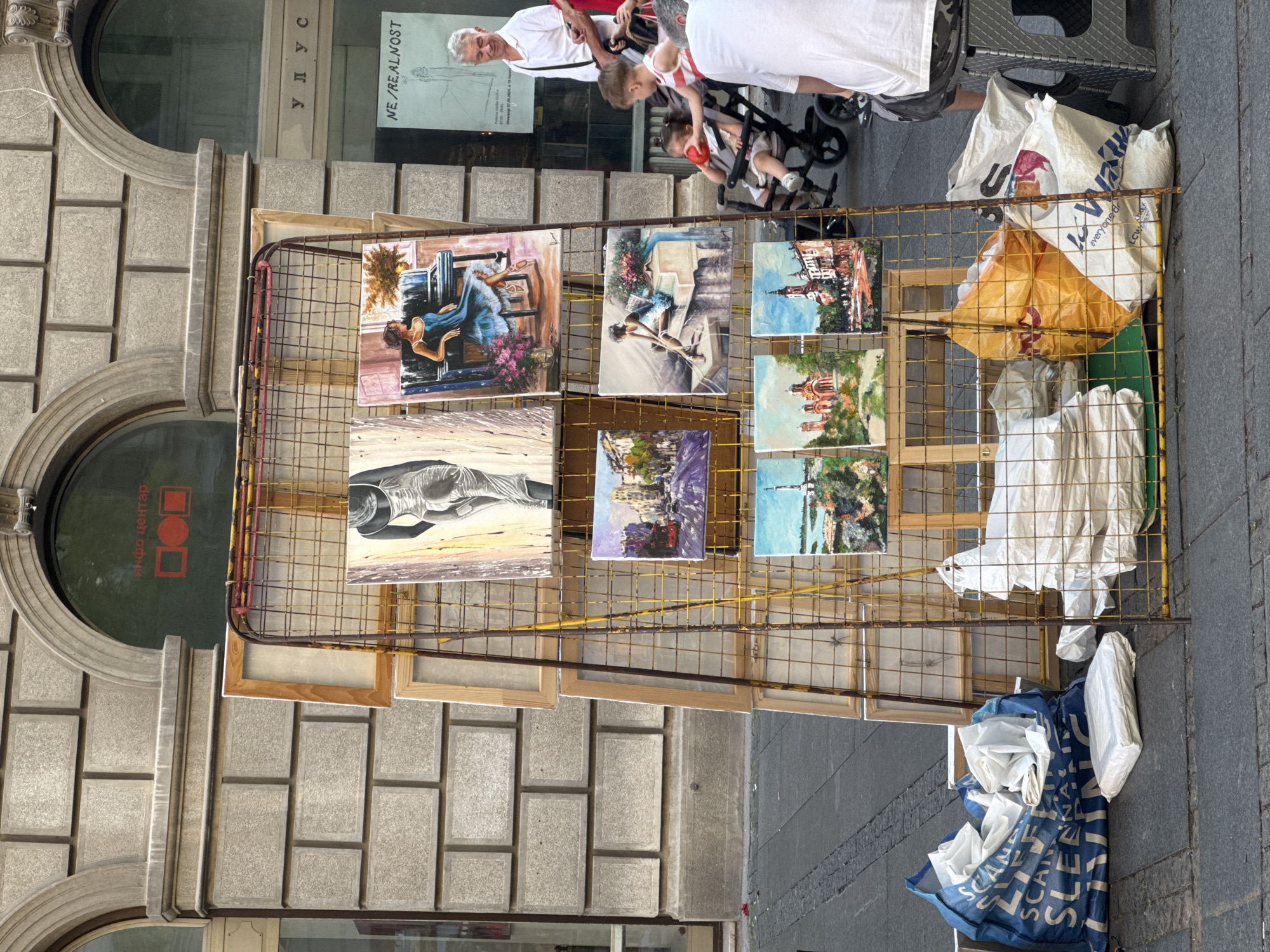 An outdoor art display is set up on a paved sidewalk in Belgrade, Serbia. A metal rack, painted yellow and rusted in places, holds several framed paintings. The paintings depict various scenes, including a woman in a dress, a woman playing a piano, cityscapes, and religious buildings. Behind the art display, a building facade with arched windows and stone block detailing is visible. One window has red lettering that appears to be Cyrillic text and a logo. Another window has the text "NE/REALNOST" and a faint drawing of a person.
In the foreground, several large bags and white packages are stacked on the ground. Some of the bags are blue and white with visible text, while others are yellow and white. To the right of the art display, a man in a white shirt and camouflage shorts is seated on a stool, facing away from the viewer. Nearby, a stroller with a young child is visible. The overall environment suggests a street market or an artist's open-air exhibition.