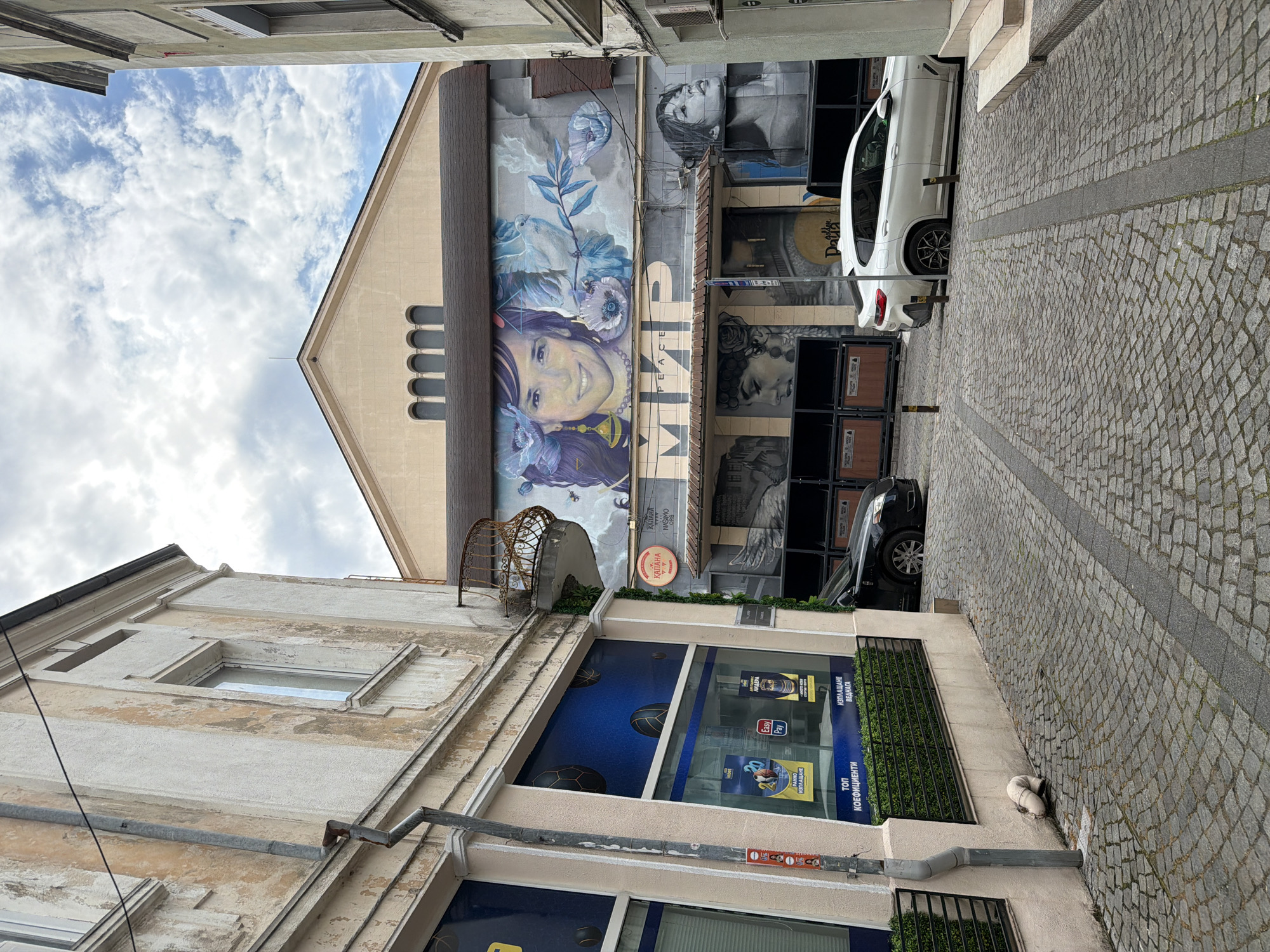 This image captures a vibrant street scene in Plovdiv, Bulgaria, likely within the Kapana district, given the sign visible. The photo is taken looking upwards through a narrow cobbled alleyway on a cloudy day.
The central focus is a large, colorful mural painted on the gable end of a building. The mural features the smiling face of a woman with long, dark hair adorned with flowers. A white dove and more blue and purple flowers are incorporated into the design. The words "PEACE" and "МИР" (the Cyrillic equivalent for 'peace') are prominently displayed below the woman's face, with "Nasimo" (likely the artist's signature) faintly visible above "МИР". The building has a light-colored facade with several arched windows near the roofline.
To the right of the main mural, on a lower section of the building facade, are black and white portraits of individuals, suggesting further artistic installations or a continuation of the mural theme. A white SUV and a dark car are parked on the cobblestone street, facing away from the viewer.
On the left side of the alley, another building with a weathered, light-colored facade houses a storefront. The shop window displays various advertisements, including "Easy Pay", "ИЗПЛАЩАНЕ ВЕДНАГА" (Immediate Payment), "ЛИДЕРА" (The Leader), and "топ КОЕФИЦИЕНТИ" (Top Coefficients/Odds), suggesting a betting or payment service. A round red sign bearing the word "КАПАНА" (Kapana) is visible above this shop. The cobblestone street features a darker stripe running down its center. An ornate, rusted metal structure extends from the left building.