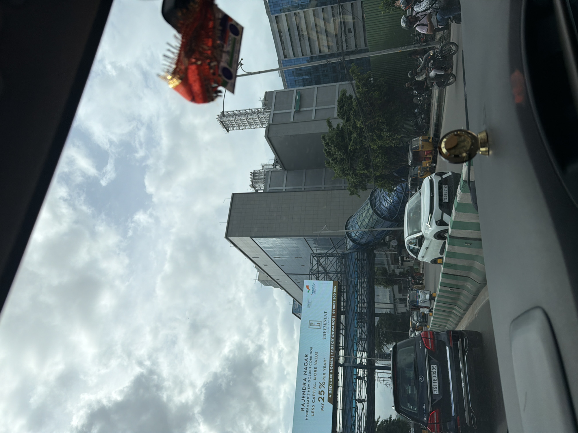 The image depicts a street scene in Hyderabad, India, likely during daytime with overcast skies. In the foreground, the dashboard and windshield of a car are visible, with a small golden emblem on the dashboard. The road ahead shows traffic, including a black Mercedes-Benz SUV with license plate TS11EJ0999, a white hatchback with license plate TG08E4169, and several auto-rickshaws. A pedestrian walkway or bridge is visible overhead, with a billboard advertising "RAJENDRA NAGAR" and "THE EMINENT," promoting residential properties with details about pricing and contact information. Behind the walkway, a large modern building complex with glass facades and concrete structures dominates the skyline. Further in the background, other buildings, trees, and a street lamp are present. A decorative red ornament with gold accents is hanging from the top right of the frame, obscuring part of the sky. The overall environment suggests an urban setting with ongoing vehicular movement.