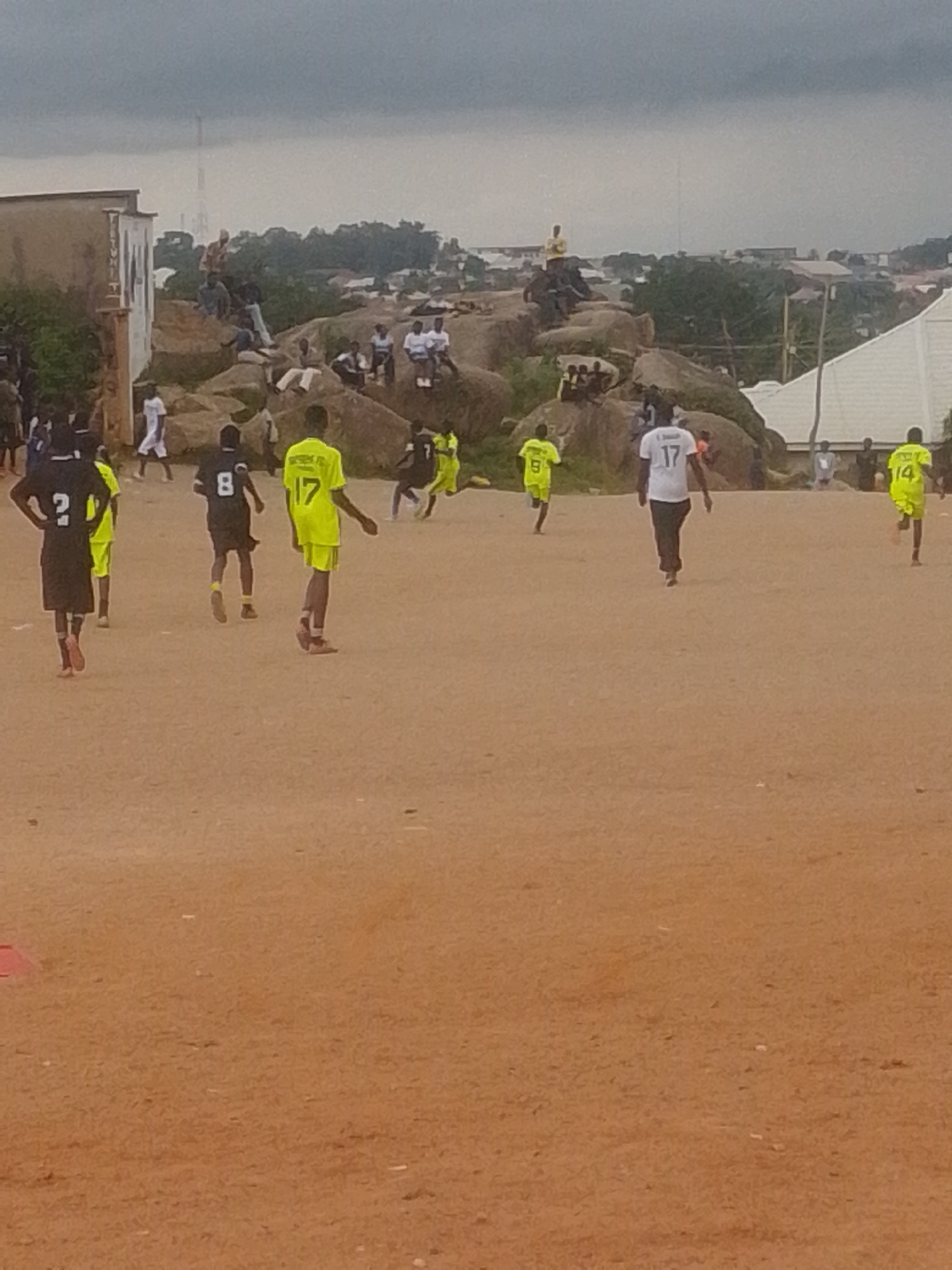 An outdoor, low-angle medium shot captures a group of young men playing soccer on a dirt field under an overcast sky. The field is sandy brown, and scattered debris is visible on its surface. In the background, a rocky outcrop with trees and buildings is situated on a hill, with a cell tower visible in the distance. A white building with a sign is to the left.
The primary subjects are the soccer players. A player in a neon yellow jersey with the number 9 on the back is running away from the camera. To his left, another player in a black jersey with the number 2 is facing away from the camera, with hands on hips. A player in a black jersey with the number 8 is also running away from the camera. Another player in a neon yellow jersey with the number 17 on the back is facing away from the camera. A player in a white jersey with the number 17 on the back is walking away from the camera. To the far right, a player in a neon yellow jersey with the number 14 on the back is running away from the camera. Several other individuals, appearing to be spectators, are scattered on the rocks in the background, some seated and some standing. The overall scene suggests a casual soccer game taking place in a somewhat undeveloped urban or semi-urban area.
