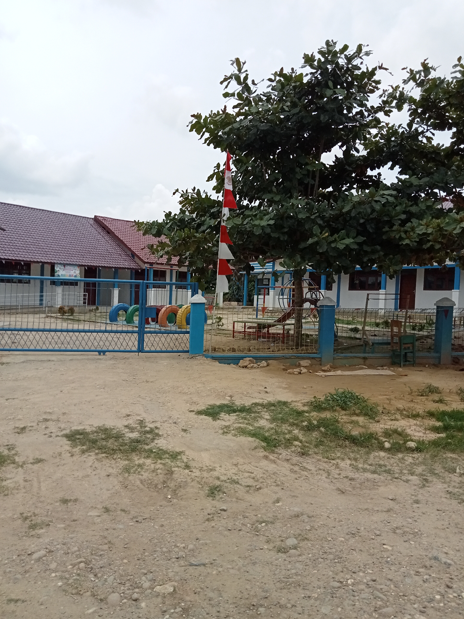 The image depicts the entrance to what appears to be a school in Langnibong, Indonesia. In the foreground, a dirt path with scattered pebbles and sparse green vegetation leads to a blue metal gate. Through the gate, a building with a red tiled roof and white walls can be seen, with windows and doors visible. Adjacent to the building, a playground area features a climbing structure and colorful tires arranged on the ground. A tall tree with dense green foliage is situated near the center of the frame, partially obscuring the building. A flag, with red and white stripes, is planted near the tree. The sky is overcast and cloudy. The overall scene suggests a tranquil, possibly rural or semi-urban environment.