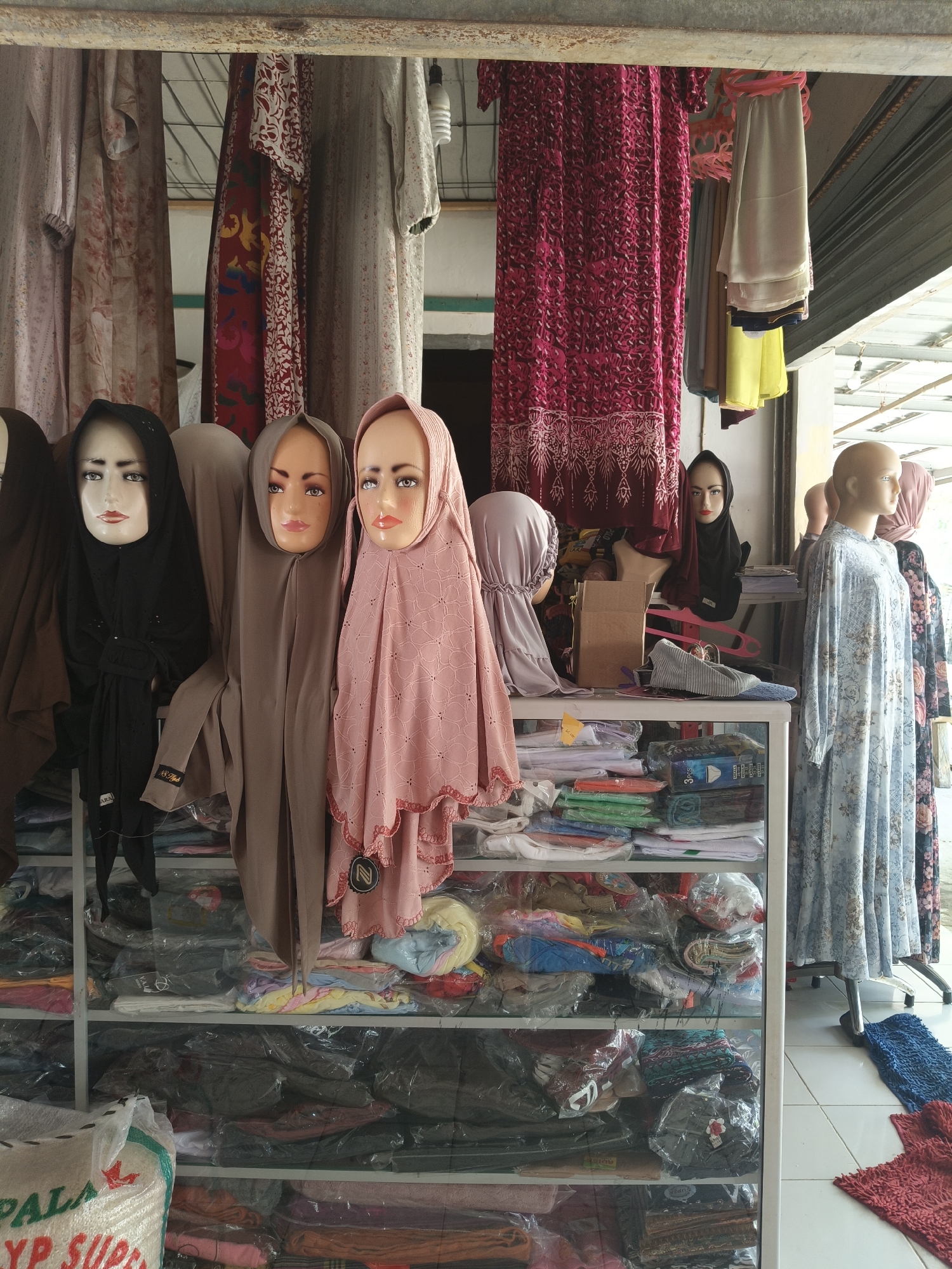 The image depicts a storefront in Rancaseneng, Indonesia, likely selling Islamic clothing and accessories. In the foreground, three female mannequins are displayed, each wearing a hijab. The mannequin on the left wears a black hijab, the middle mannequin a taupe hijab, and the mannequin on the right a pink hijab. Behind them, various garments, including dresses and headscarves, hang from a rack. A shelf unit filled with neatly folded and packaged clothing items is visible below. Further back, other mannequins and displays of clothing can be seen. The storefront appears to be an outdoor or semi-open space, with a corrugated metal roof overhead. The overall impression is of a retail environment specializing in modest women's apparel.
