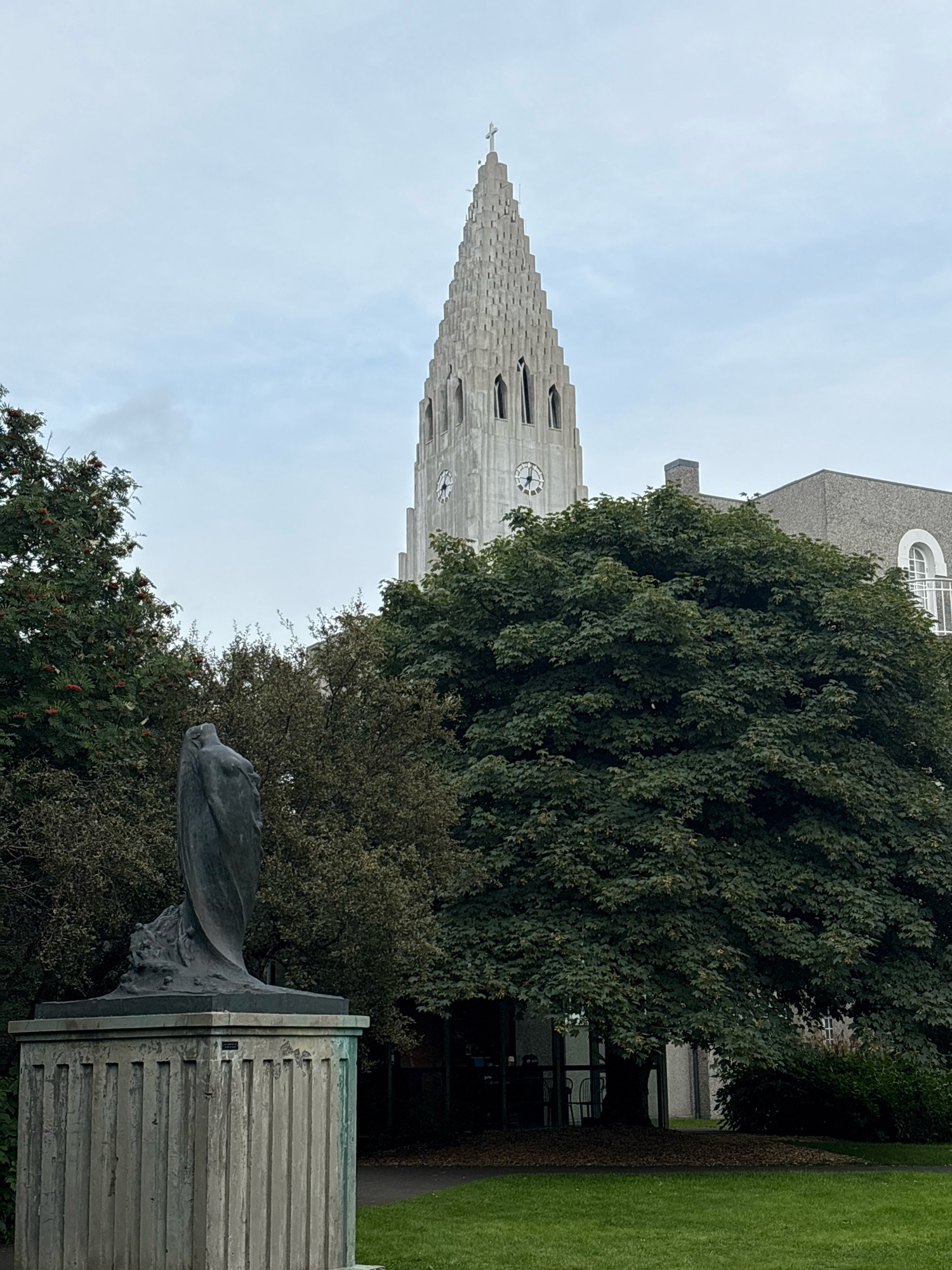 The image shows Hallgrímskirkja church in Reykjavik, Iceland, as seen from a park. In the foreground, a dark bronze statue of a woman is mounted on a weathered concrete pedestal. The statue depicts a figure with her arms clasped in front of her, with what appears to be a wave-like base. Behind the statue, a large, leafy green tree dominates the mid-ground. Hallgrímskirkja's distinctive tower rises above the trees in the background, with its patterned concrete facade and a clock visible. The sky is overcast and pale blue. To the right of the church tower, part of another building with a textured gray facade and arched windows is visible. The ground in the foreground is a neatly mown green lawn.