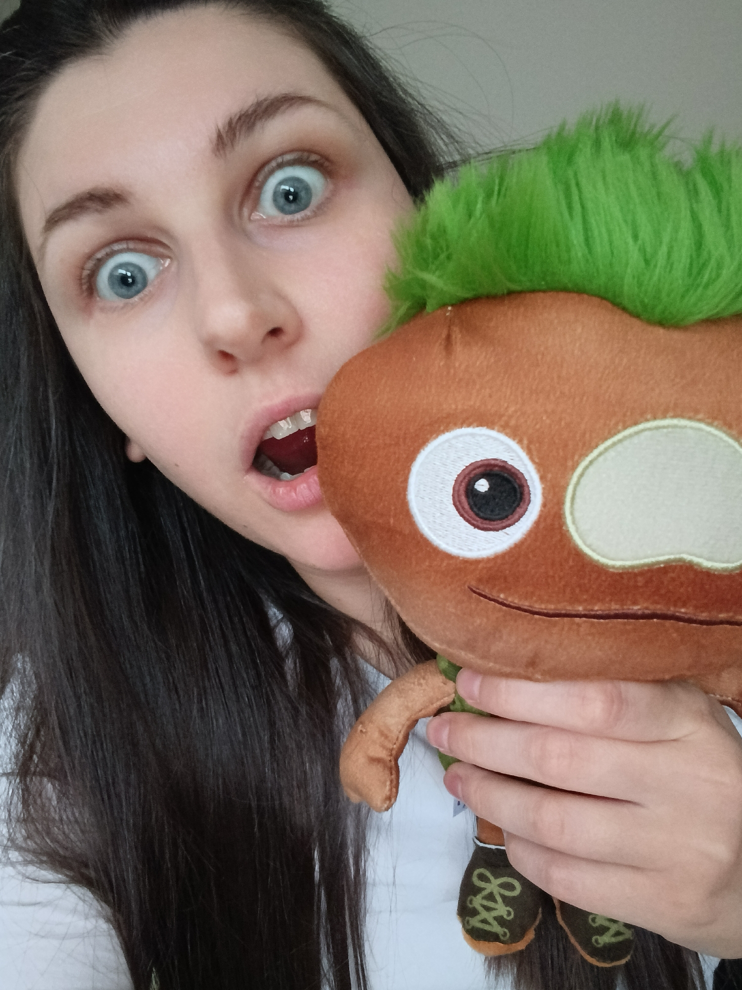 A medium-shot photograph shows a fair-skinned woman with long, dark hair holding a plush toy. The woman's eyes are wide, and her mouth is open in a surprised expression. The plush toy is brown with green "hair" on top, and has embroidered features including eyes and a smile. The toy appears to be wearing shoes. The woman is wearing a white top. The background is not visible. The location is Unknown, Unknown.