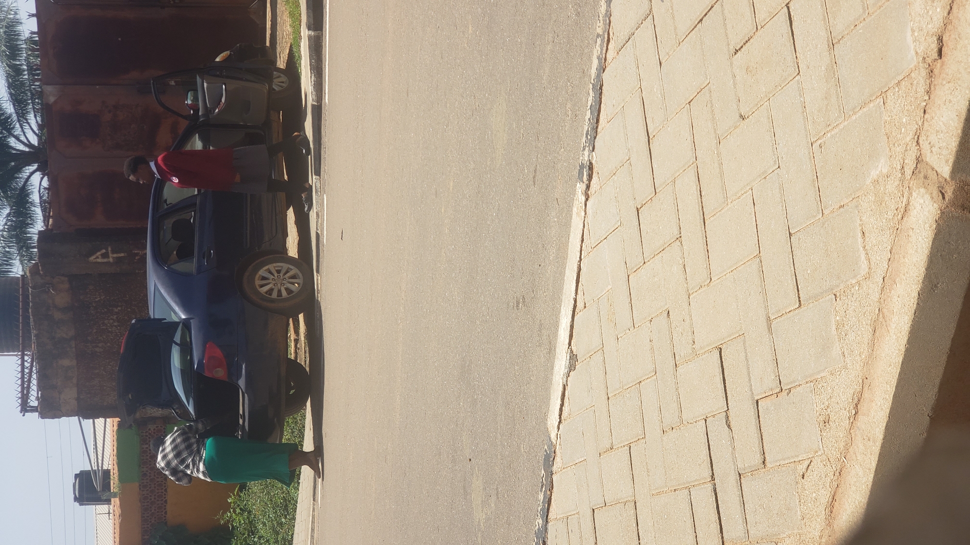 In Narayi, Nigeria, a dark blue sedan with its trunk open is parked on the side of a paved road. Two individuals are near the car. To the left, a person wearing a checkered shirt and a long green skirt is reaching into the open trunk. To the right, a second person, dressed in a red jacket over a white shirt and a gray skirt, stands beside the open passenger door of the car. The environment appears to be daytime with clear skies. The road has a cobblestone sidewalk adjacent to it. In the background, there are buildings and a large metal gate.