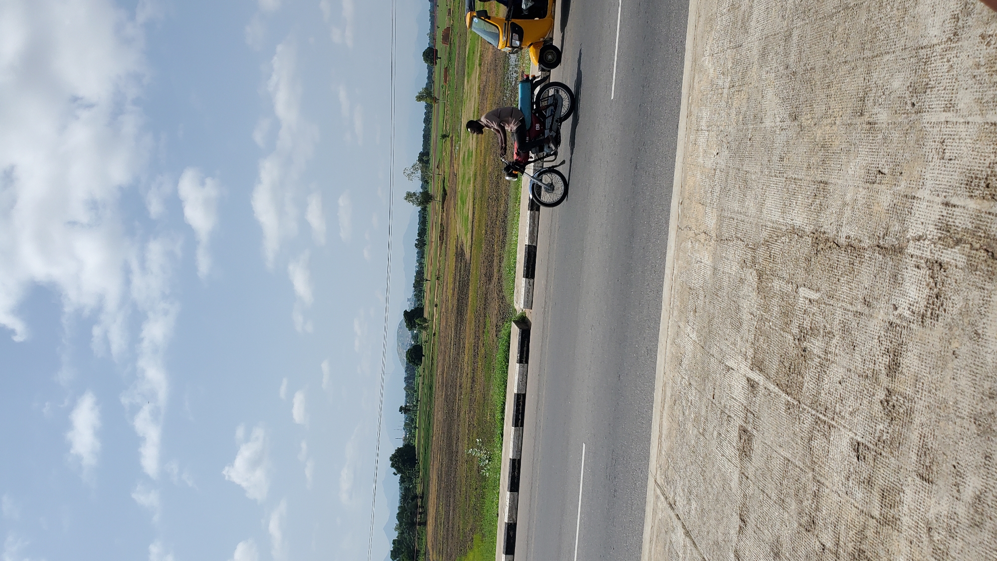 A person is riding a motorcycle on a paved road in Jalingo, Nigeria. The rider, wearing a brown shirt and red pants, is positioned in the center of the frame, with their body leaning forward. The motorcycle is a dark color with silver accents and has a black seat. To the right of the motorcycle, a yellow auto-rickshaw is parked. The road has a white line marking the lane and a striped black and white border.
The background features a vast, cultivated field with patches of green and brown, suggesting agricultural activity. In the distance, a mountain range is visible under a bright, partly cloudy sky, indicating it is daytime with fair weather. The overall scene depicts a rural or semi-rural environment. The concrete structure in the foreground on the right side of the image is textured and appears to be part of a bridge or overpass. No visible text is present in the image.