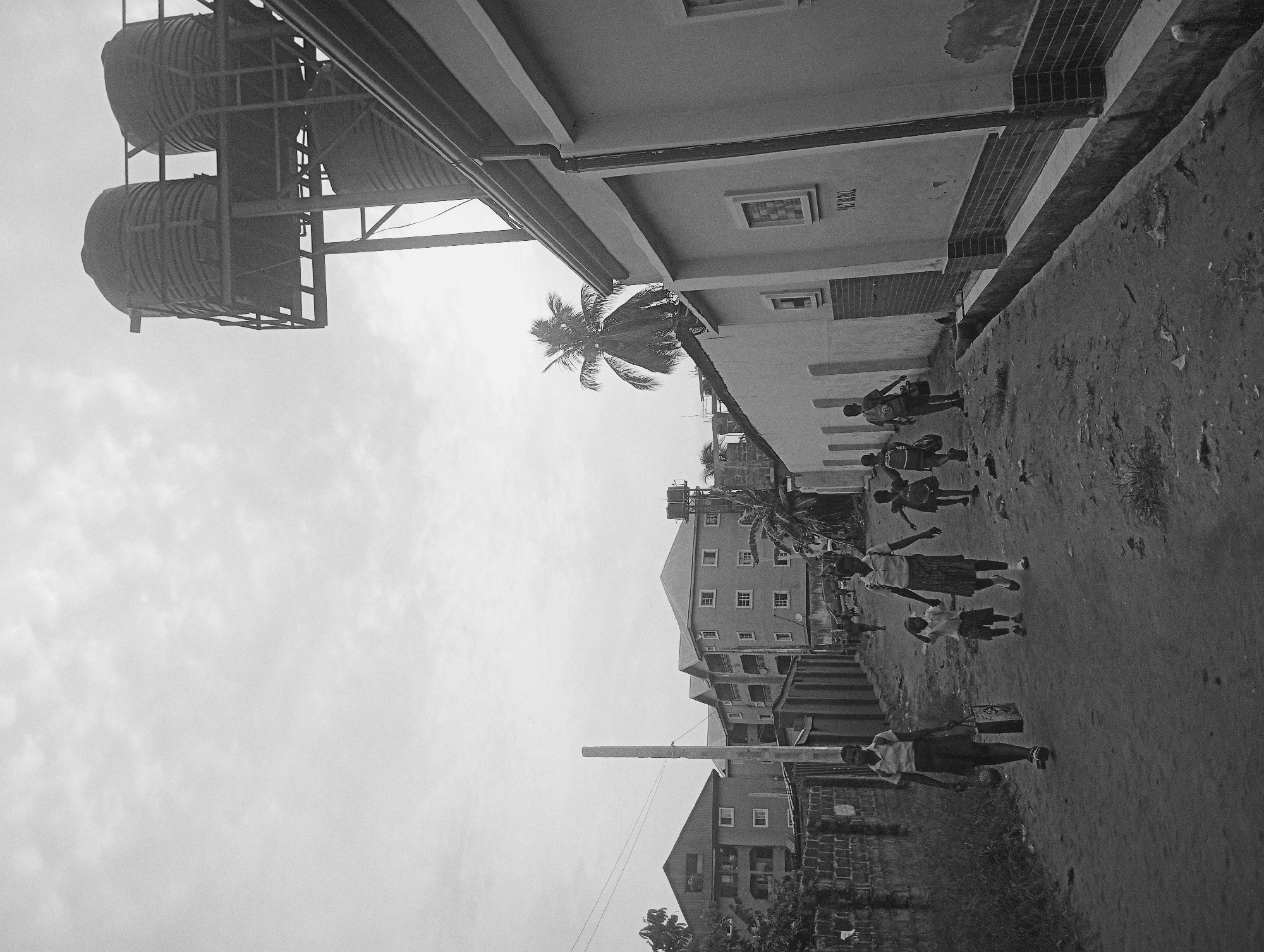 A black and white image depicts a street scene in Nkpor, Nigeria. Several children, dressed in school uniforms, walk along a dirt road. A palm tree leans towards them. Adjacent to the road is a building with the word "PETROL" on it. A tower with water tanks sits atop a building on the left side of the image. Further down the road are residential buildings. The sky is overcast.