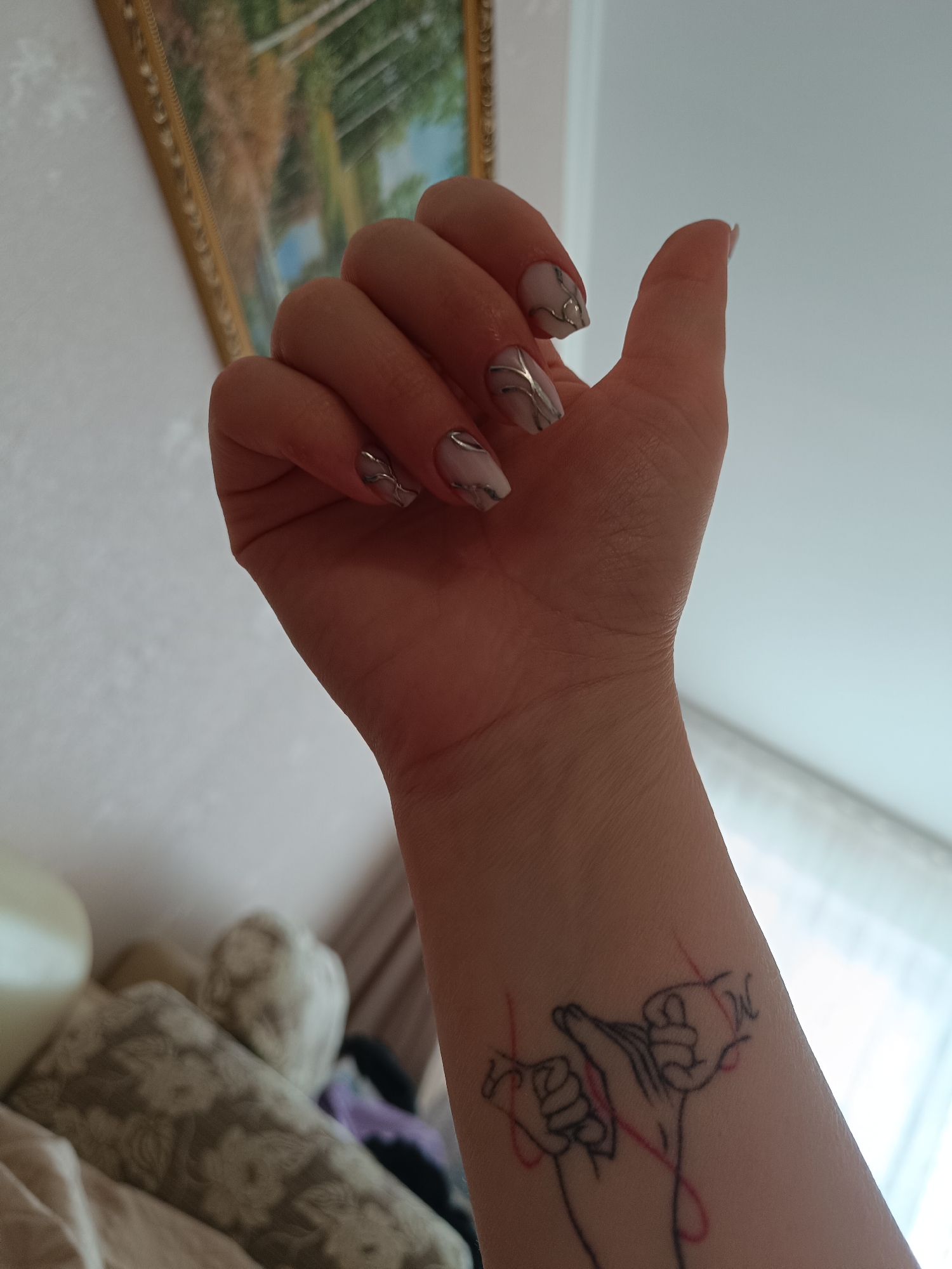 A right arm and hand are prominently featured in the image. The fingernails are polished with a light-colored base and silver vein-like accents. A tattoo is visible on the forearm, depicting two hands connected by a red line. The background includes a framed artwork and soft furnishings. The location is not identifiable as Tobolsk, Russia from the image provided.