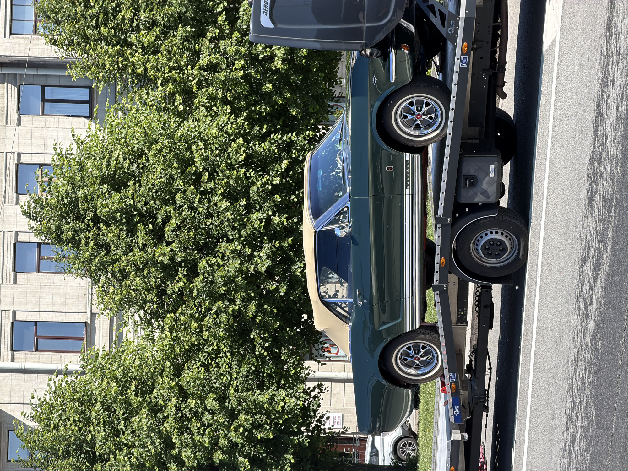 A dark green 1960s Ford Mustang convertible with a tan soft top is parked on a flatbed trailer. The car has chrome accents, whitewall tires, and classic wire-spoke wheels. The trailer is attached to a dark gray truck cab, which is partially visible on the right side of the frame. The truck and trailer are on a paved road, with a tree-lined sidewalk and a building in the background. The building is light-colored with multiple windows. The scene appears to be in Saint Petersburg, Russia, given the architectural style of the building and the presence of a Cyrillic sign on the sidewalk. The lighting suggests it is daytime.