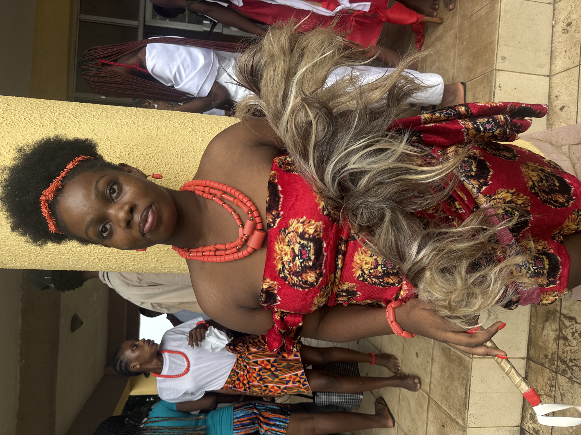 The image captures a portrait of a young woman in traditional attire, set against a backdrop that suggests an outdoor event or gathering in Ihiagwa, Nigeria. The woman is adorned with a vibrant red and gold patterned wrap dress and multiple strands of coral bead necklaces and bracelets. A coral-colored headband with a matching earring is also visible. Her hair is styled with a blonde wig that cascades down her back. She holds a decorative staff-like object in her right hand.
In the background, other individuals are partially visible, also dressed in traditional clothing. One woman in the middle ground wears a white top and a patterned skirt, with coral beads around her neck. Another person in the background is seen in a white garment with long red braids. The ground is paved with light-colored tiles, and a yellow wall runs along the left side of the frame.
The scene appears to be from a daytime event, given the natural lighting. The overall mood is festive and celebratory, suggested by the colorful attire and the communal gathering. The image focuses on the woman in the foreground, highlighting her striking appearance and cultural regalia.