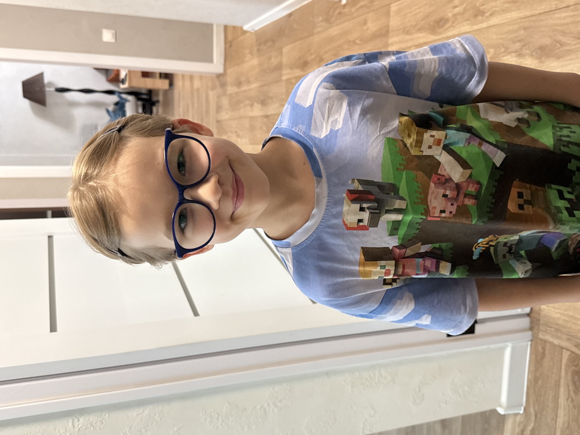 A child with short blond hair and blue glasses is smiling at the camera. The child is wearing a blue and white patterned shirt with a Minecraft print. The background includes a white wall, a white doorway, and a wooden floor. The location of the image is not ascertainable based on the context.