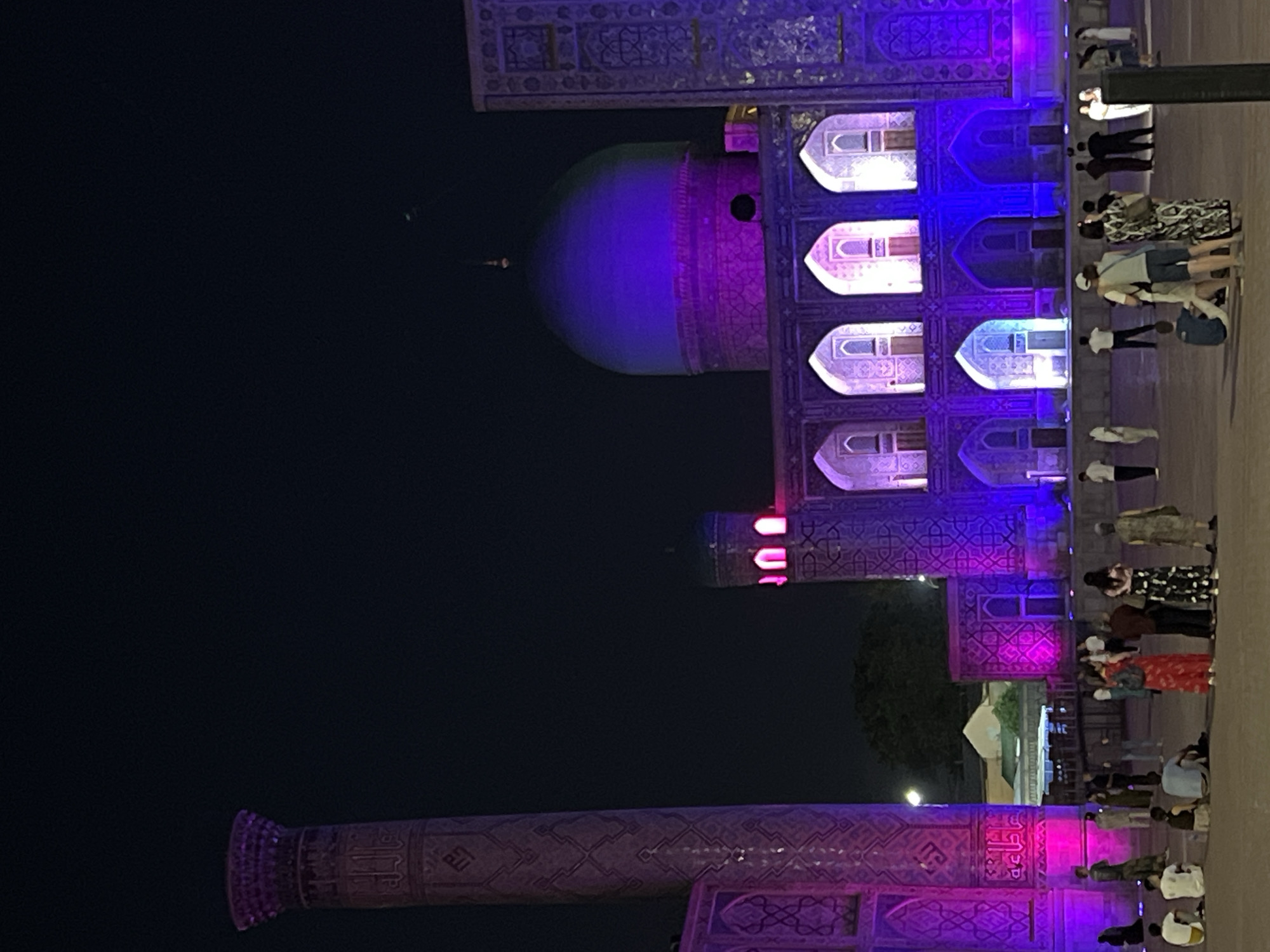 This media file depicts an outdoor scene at night, likely in Samarkand, Uzbekistan, given the architectural style and lighting. The central focus is a large, ornate structure illuminated by vibrant purple and blue lights. This structure appears to be a historical monument or building, characterized by intricate geometric patterns, arched windows, and a prominent dome.
A group of people are visible in the foreground and mid-ground, observing the illuminated building. They are casually dressed, suggesting a tourist or evening outing. Their presence indicates that an event or attraction is taking place, drawing people to this location.
The time of day is clearly night, as evidenced by the dark sky and the artificial lighting on the structure. The lighting creates a dramatic and enchanting atmosphere, highlighting the detailed craftsmanship of the architecture. There are no discernible weather cues, but the dark sky suggests clear conditions.
Notable details include the rich ornamentation of the building, the contrast between the dark sky and the bright lights, and the diverse attire of the individuals. The overall emotion evoked is one of awe and wonder, likely inspired by the beauty of the illuminated monument. There is no visible text in the image.