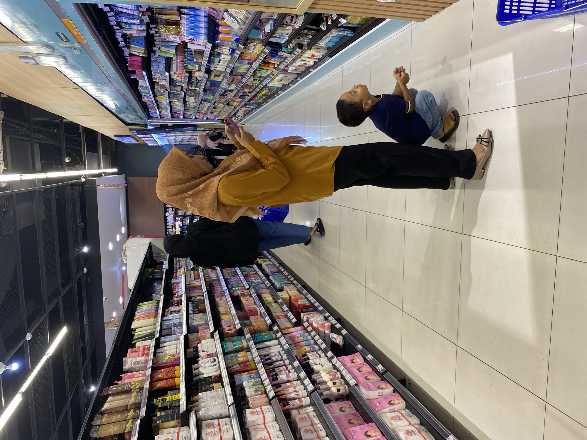 In a brightly lit supermarket aisle in Banda Aceh, Indonesia, a young boy wearing a navy blue t-shirt and blue jeans squats down to look at products on a lower shelf. To his left, a woman in an orange headscarf and yellow long-sleeved shirt, also wearing black pants, stands and uses her mobile phone. Behind them, further down the aisle and partially obscured, a person in a black hijab and blue jeans walks away from the camera. The shelves on both sides of the aisle are stocked with various personal care products, including bottles of lotion, shampoo, and packaged items. The floor is tiled in a light neutral color. To the right, a blue shopping cart is partially visible. The scene suggests a typical shopping outing.