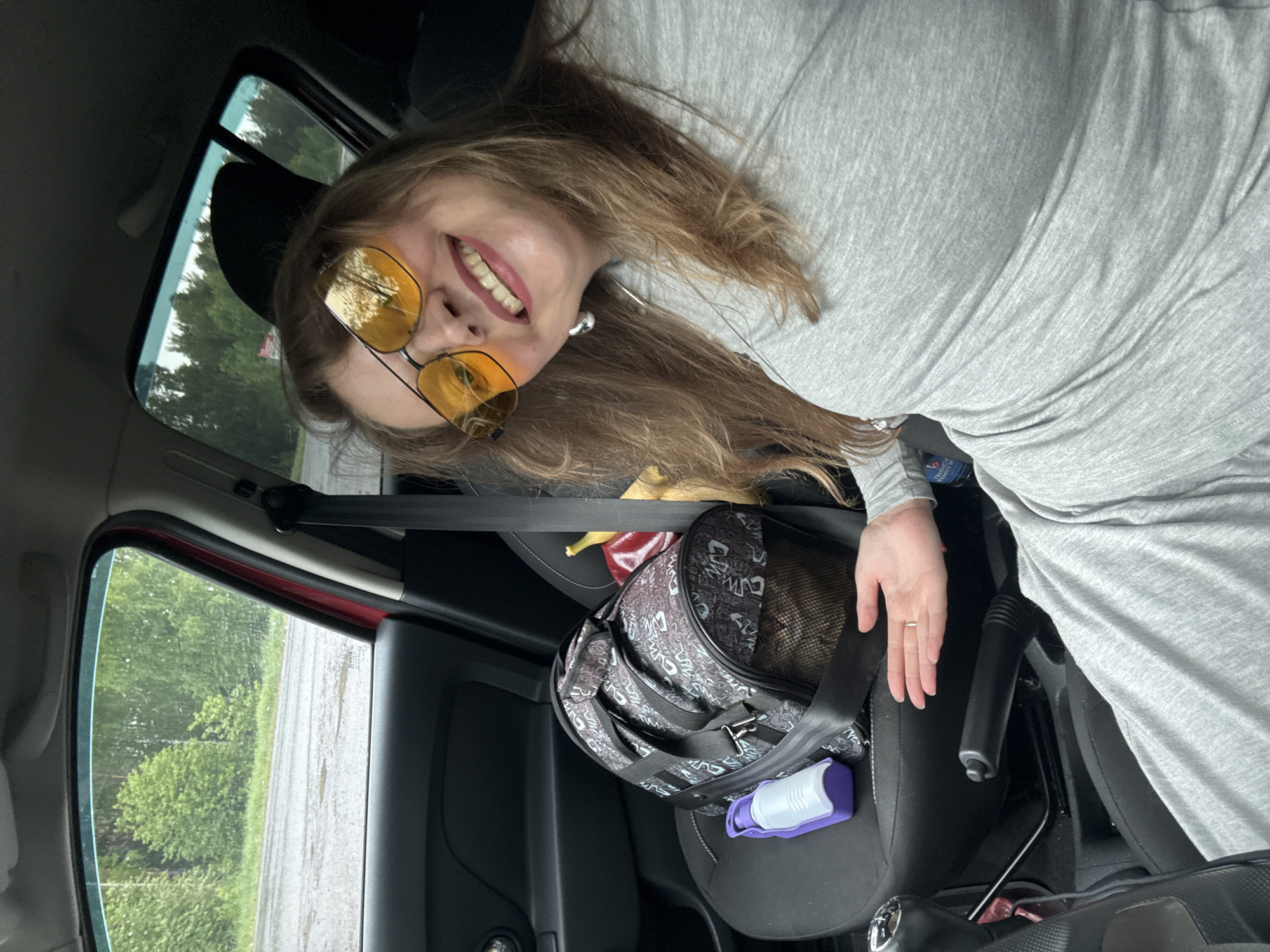 A woman with long blonde hair and yellow-tinted glasses is reclining in the front passenger seat of a car, smiling towards the camera. She is wearing a grey long-sleeved shirt. A patterned pet carrier is secured on the passenger seat with a seatbelt. Inside the carrier, an animal is partially visible. A purple and white object is attached to the side of the carrier. A car window shows a view of trees and possibly a wet road. The image was most likely taken in the city of Mozhga, Russia.