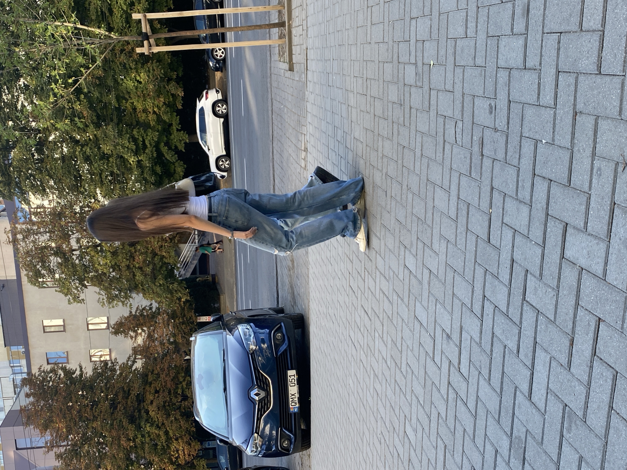 An outdoor, full-shot, eye-level, vertical photograph depicts a woman walking on a brick sidewalk in Chișinău, Moldova. She is seen from the back and her long dark hair extends to her waist. She wears a white tank top and light blue, distressed, wide-leg jeans. Her left foot is slightly raised as she walks. A white purse with a black strap is slung over her left shoulder.
A dark blue Renault Scenic is parked on the street to the left, displaying a Moldovan license plate that reads "QMX 051". To the right of the blue car, a white SUV and a black sedan are parked on the same street. Beyond the street, trees with autumnal foliage are visible, and a light-colored building with windows is in the background. The sun casts shadows on the ground, indicating it is likely late afternoon. The brickwork of the sidewalk is laid in a herringbone pattern.