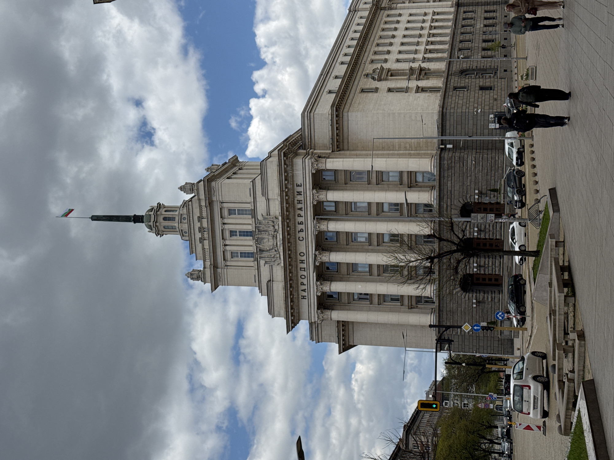 This image captures a daytime scene in Sofia, Bulgaria, featuring the prominent National Assembly building. The main subject is a grand, multi-story building constructed in a classical architectural style with large columns, ornate cornices, and numerous windows. A distinctive central spire, topped with the Bulgarian flag (horizontal white, green, and red stripes), rises above the building. The facade is light-colored, possibly stone or concrete.
Clearly visible text in Cyrillic characters on the building reads "НАРОДНО СЪБРАНИЕ," which translates to "National Assembly," confirming the building's identity as the Bulgarian Parliament.
On the paved sidewalk adjacent to the building, several people are seen walking. Some are dressed in everyday clothing. Along the street, various cars are parked, including a white hatchback and darker sedans. Bare trees suggest either late winter or early spring. Traffic signs and a green traffic light are visible in the distance, indicating an active urban environment.
The sky is partially cloudy, with patches of bright blue sky interspersed with white and grey clouds, suggesting fair weather. The lighting indicates it is daytime, likely morning or afternoon. The overall scene depicts a typical day around a significant landmark in Sofia.