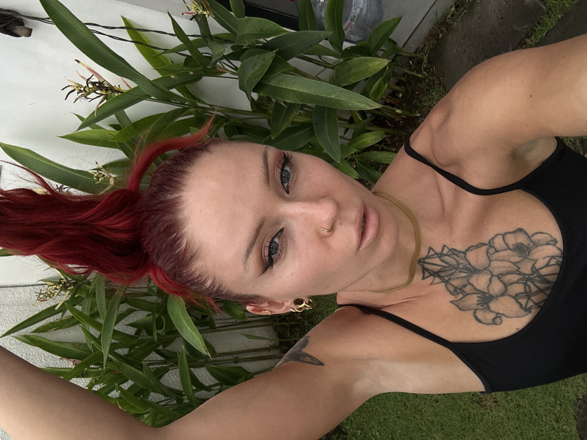 A selfie shows a woman with bright red hair pulled into a messy bun. She is wearing a black tank top and a gold necklace. A large floral tattoo is visible on her chest. She has dramatic winged eyeliner and a nose ring. The background is filled with lush green plants and a white wall. The image appears to have been taken outdoors, possibly in a garden setting in North Kuta, Indonesia.