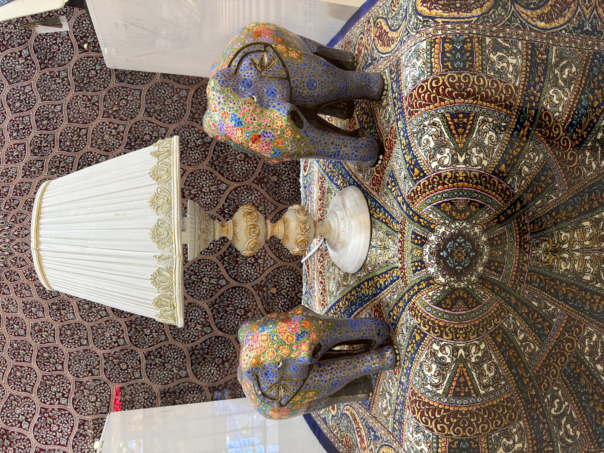 The image depicts an interior setting, possibly in Baan Chaweng, Thailand. On the left is a lamp with a white, pleated shade trimmed with gold. Its ornate base is primarily cream-colored with gold embellishments. Two decorative elephant statues with blue bodies and colorful floral designs adorn the scene. The figures are placed atop a patterned carpet. The floor covering features a complex design, displaying shades of red, blue, gold, and brown. A section of the floor or wall, patterned in repeating motifs, is visible to the left of the lamp.