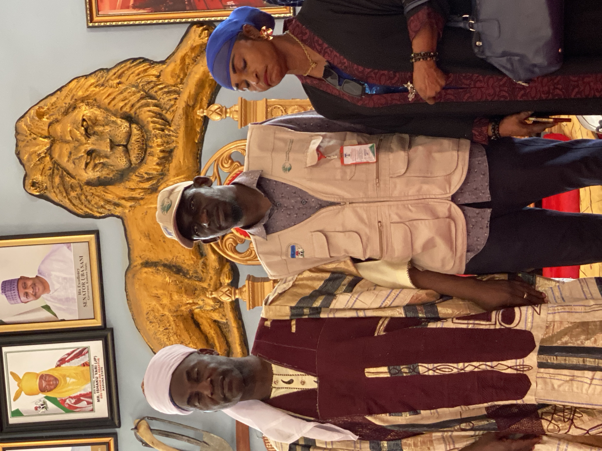 The image depicts three individuals in an indoor setting, likely in Sabon Tasha, Nigeria, given the signage in the background. The person at the bottom is dressed in a traditional robe. The man in the center is wearing a vest. The woman at the top is adorned in a dress and a blue headscarf. Behind them, a gold lion statue is visible, as well as framed portraits with the text "HIS ROYAL HIGHNESS MR. ISHAKU YARI (JP) ESU CHIKUN (SA GBAGYIR KADUNA STATE" and "His Excellency SENATOR UBA SANI Governor, Kaduna State.".