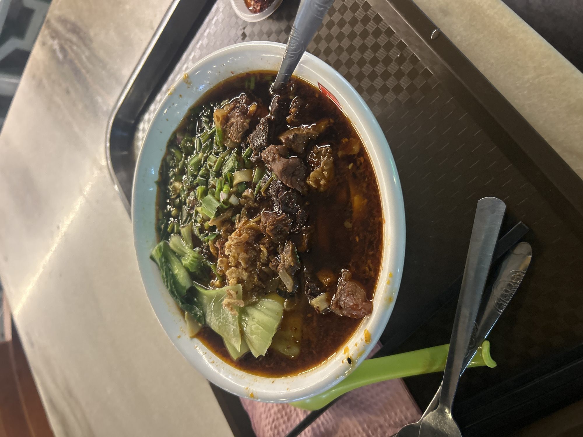 A close-up, high-angle shot shows a white bowl filled with a dark broth, noodles, chunks of beef, and green vegetables, possibly bok choy, and chopped scallions. The bowl is placed on a brown plastic tray with a checkered pattern. To the left of the bowl, a lime green utensil handle is visible, as well as two metal spoons and chopsticks. A small red dish containing a chili paste is positioned to the right of the bowl on the tray. The background consists of a table with a beige surface and a darker edge, likely the surface of a restaurant table in Ipoh, Malaysia. The lighting suggests an indoor setting, possibly a hawker stall or a casual eatery.