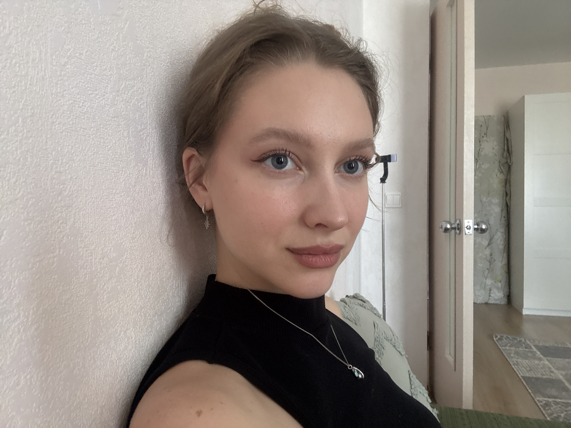 A young woman with blonde hair pulled back and blue eyes poses for a selfie. She is wearing a black, sleeveless turtleneck shirt and a silver necklace with a pendant. Her left ear is adorned with a small, star-shaped earring. She is leaning against a textured white wall, with her right shoulder visible in the foreground. In the background, a partially open door reveals a hallway with a patterned wallpaper and a rug on the floor. A phone on a tripod is visible to the right of her head. The scene appears to be an indoor residential setting in Izhevsk, Russia.
