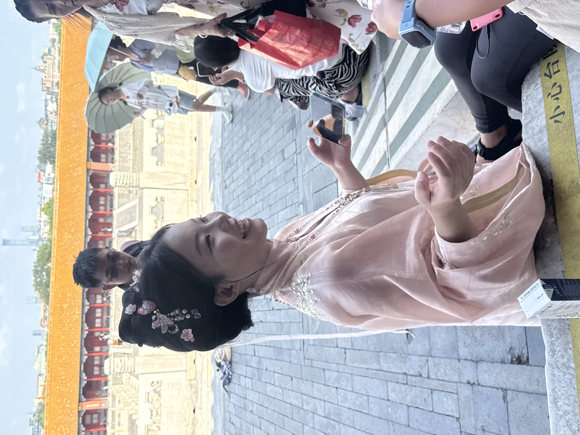 In Dongcheng District, China, a young woman in traditional attire, adorned with flowers and pearls, smiles broadly while holding a smartphone. Her pastel pink robe drapes over her shoulders, and her hair is styled elaborately. Behind her, a man in a dark jacket and glasses is visible. In the background, people with umbrellas stroll past traditional Chinese architecture with orange tiled roofs, and modern city buildings can be seen in the distance. The scene takes place on stone steps, with a yellow caution sign on the lower step. Other individuals are also present, some holding bags and phones, indicating a public space likely frequented by tourists.