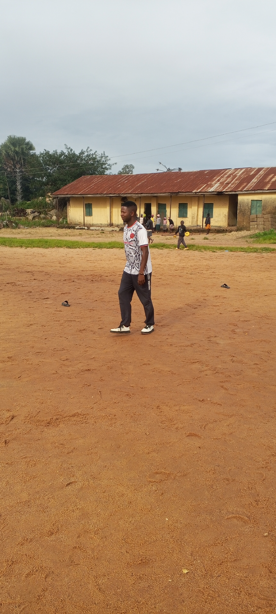 A young Black man, wearing a white t-shirt with a black and red pattern and dark gray pants, walks across a dirt field. He is facing right and looking to his left. In the background, there is a long, single-story building with a rusty, corrugated metal roof and yellow walls. Several other people, including children, are visible near the building. There are trees and vegetation behind the building, and the sky is overcast. The location is identified as a school in Ohit, Nigeria.
