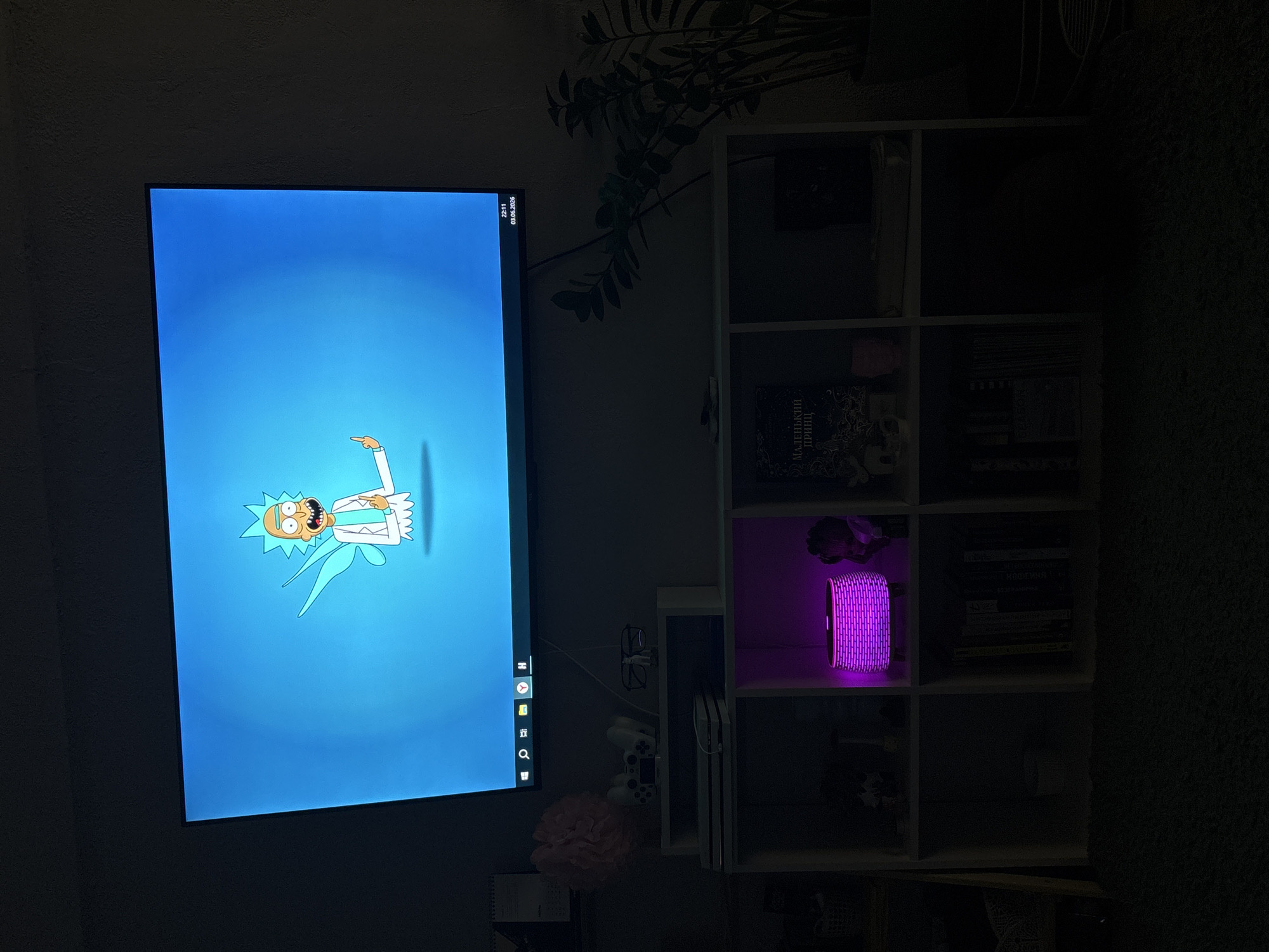 The dimly lit indoor scene captures a corner of a room, likely a living room or bedroom, dominated by a large television screen on the left and a modular cube-style shelving unit on the right. The subdued lighting suggests late evening or night.
The television screen displays a vibrant light blue background with a stylized cartoon character, Rick from Rick and Morty, positioned in the center, pointing to the right. A taskbar with various application icons is visible at the bottom of the screen. In the top right corner of the screen, the time "22:11" and the date "03.03.2025" are displayed.
To the right of the TV, a white modular shelving unit is filled with various items. In one of the middle-left cubes, the spine of a book titled "МАЛЕНЬКИЙ ПРИНЦ" (The Little Prince in Russian) is visible. Adjacent to it, in the middle-right cube, a cylindrical object emits a striking purple glow, suggesting it is an illuminated lamp, smart speaker, or air purifier. Further down, a white PlayStation console (likely a PS4 or PS5) is placed horizontally in one of the lower cubes, with a white controller nearby.
Behind the TV and partially draped over its top right corner, a dark-leaved houseplant (possibly a ZZ plant) adds a touch of greenery. Other details include a pair of glasses hanging from a wire near the TV, a pink ruffled decorative item below the TV, and a small notebook or planner on a surface beneath it. No specific activity is taking place; the scene is a static interior shot. The visible Russian text on the book aligns with the provided location context of Zheleznogorsk, Russia.