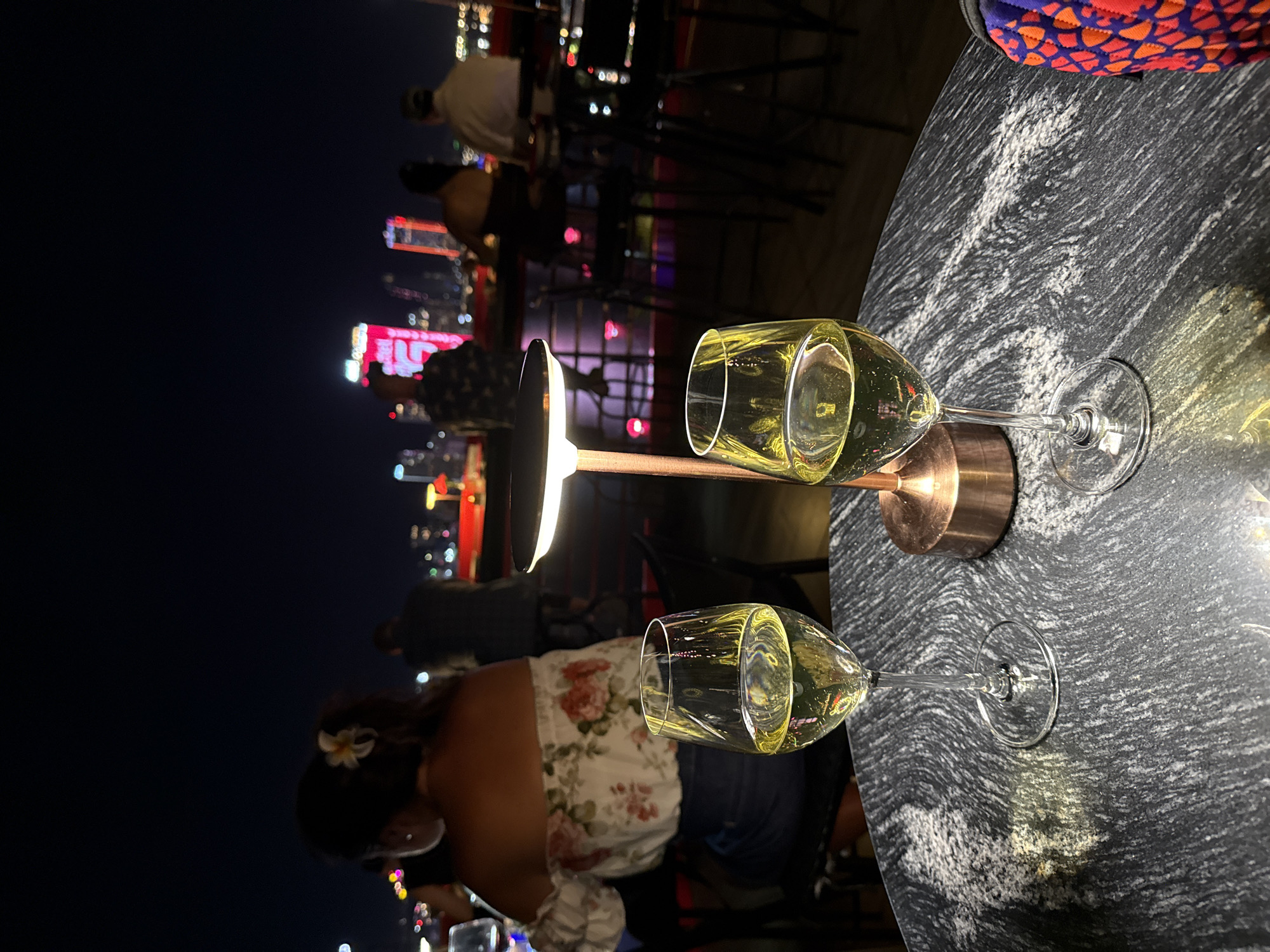 This image depicts an evening scene at a rooftop establishment in Da Nang, Vietnam, offering a view of the city skyline.
In the foreground, two stemmed glasses containing a pale yellow liquid, consistent with white wine, rest on a dark grey stone tabletop with distinct white veining. Between the glasses, a modern, minimalist table lamp with a circular light source and a copper-colored cylindrical base illuminates the table. To the right of the table, a partial view of an object covered in a vibrant fabric, featuring a pattern of red-orange scales on a purple background, is visible.
In the mid-ground, several individuals are present, out of focus. On the left, a woman is seen from behind, wearing an off-the-shoulder white top adorned with a floral pattern of pink/red blossoms and green foliage. A white plumeria flower is tucked into her dark hair, which is styled in a ponytail or bun. A large, silver-toned hoop earring is visible on her left ear. Other patrons are indistinctly visible further back, seated at tables or standing, against the backdrop of the city.
The background reveals the illuminated cityscape of Da Nang at night under a dark sky. Two prominent high-rise buildings are distinctively lit in red. The building on the left clearly displays the acronym "ACV" near its peak, identifying it as the Da Nang Administrative Center. Other blurred lights and structures suggest a dense urban environment. The overall scene captures a relaxed atmosphere at an outdoor venue with an urban vista.