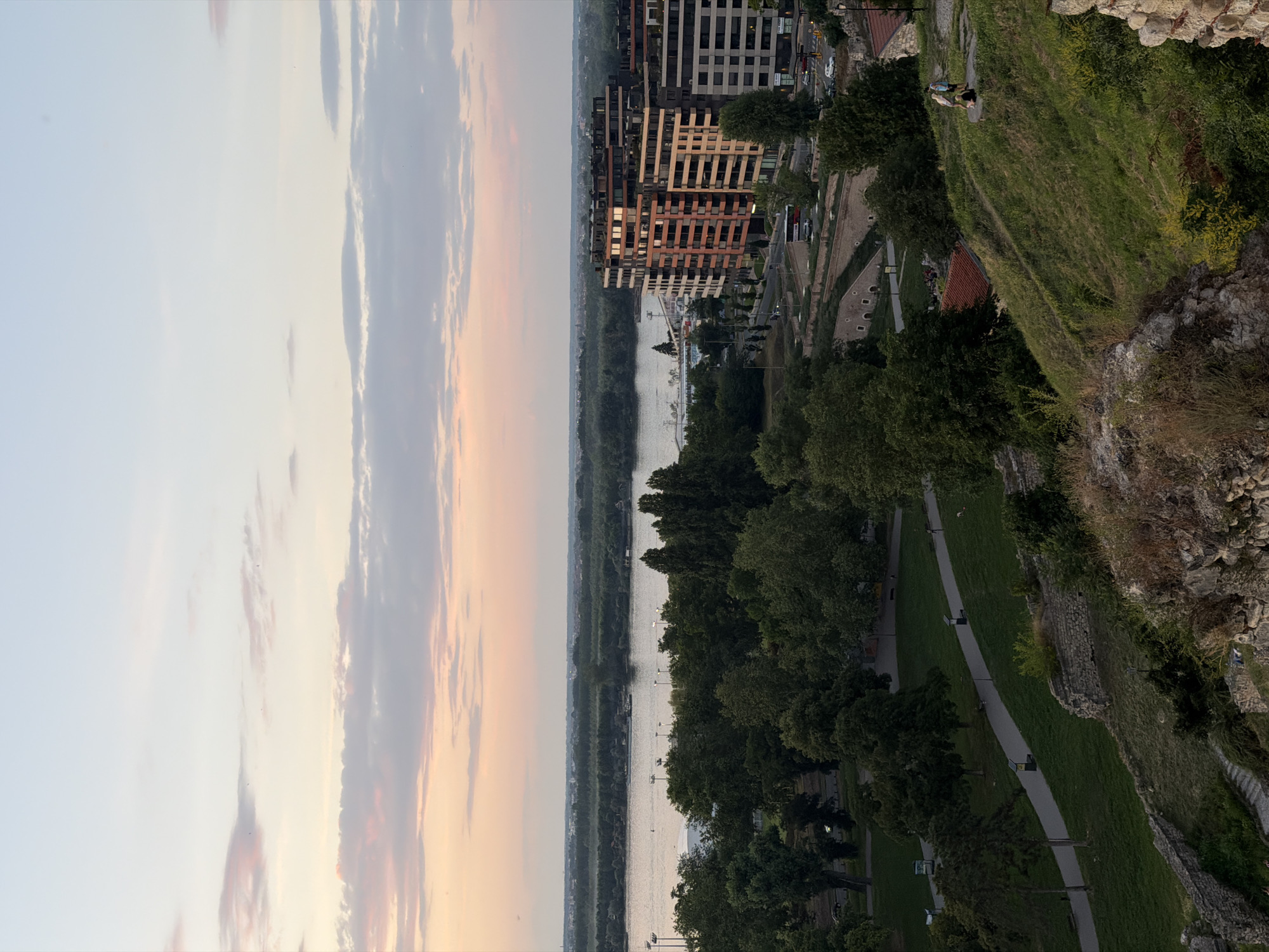 The image depicts a panoramic view of Belgrade, Serbia, likely taken at dusk from a high vantage point. In the foreground, a grassy slope is dotted with rocks and vegetation, suggesting a park or historical site. A winding paved path leads through the green expanse, with a few individuals visible walking or standing.
Beyond the immediate foreground, a dense line of trees forms a verdant band, separating the park area from a wide river, which occupies the middle ground of the image. A barge or small ship is present on the water. Across the river, a distant, tree-covered landscape stretches towards the horizon, with hints of urban development visible.
In the right middle ground, modern multi-story buildings with balconies and patterned facades stand prominently. These buildings suggest a contemporary urban development adjacent to the park and river. Further in the distance, the sky is painted with soft hues of pink, orange, and blue, characteristic of a sunset. Wispy clouds are scattered across the sky, adding to the serene atmosphere.
The overall scene captures a blend of natural beauty and urban architecture, with the river acting as a central feature connecting the different elements of the city. The lighting suggests it is late afternoon or early evening, with the sun casting a warm glow over the landscape.