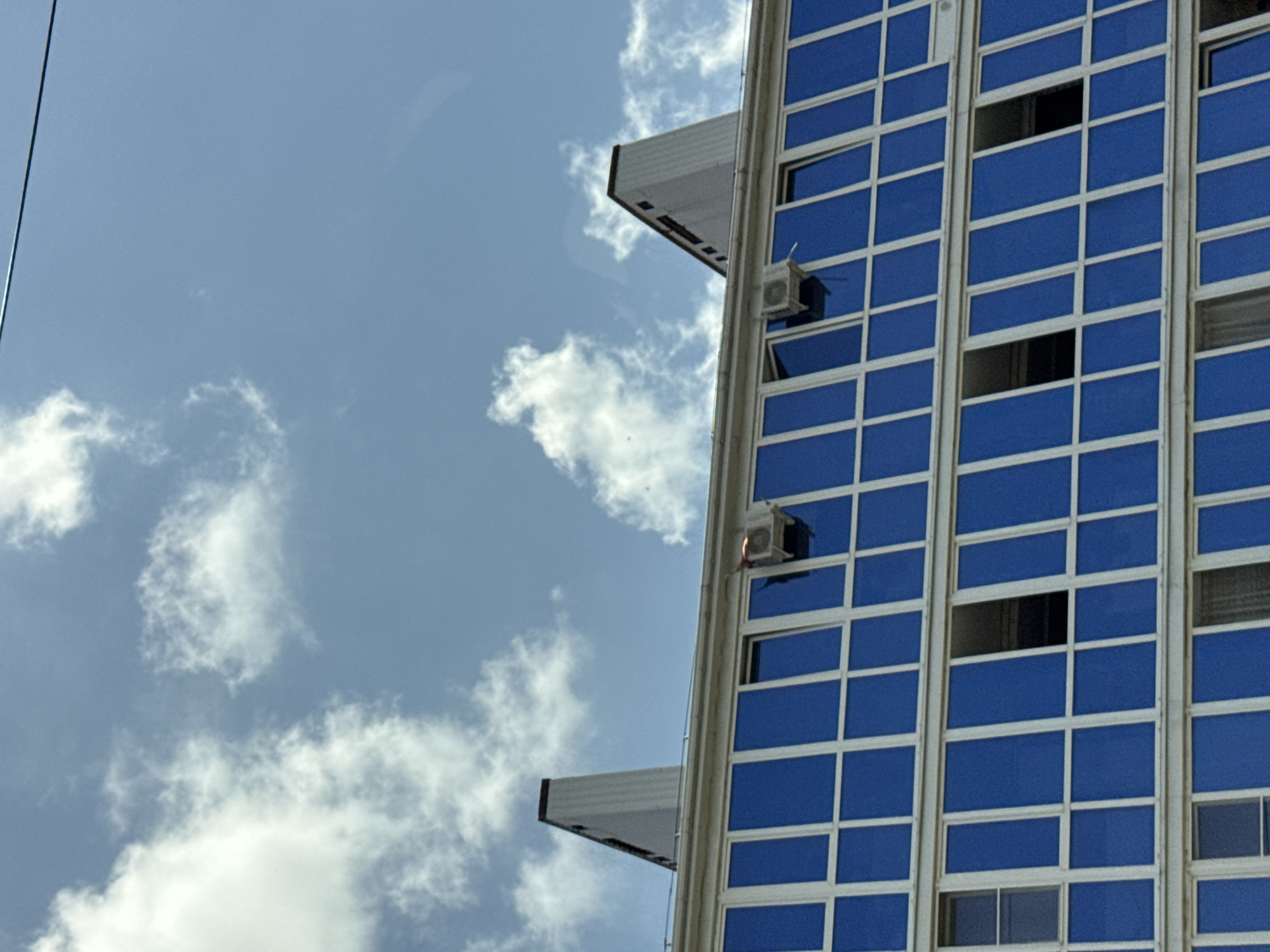 The image captures the upper facade of a multi-story building with striking blue windows, set against a bright, cloudy sky. Two prominent white architectural elements, resembling elongated pyramids or spires, rise from the building's roofline, one on the left and another on the right, angled against the sky. Visible on the building's exterior are two air conditioning units. The building appears to be located in Mikhaylovsk, Russia, as indicated by the provided context. The overall scene suggests an urban environment during daylight.