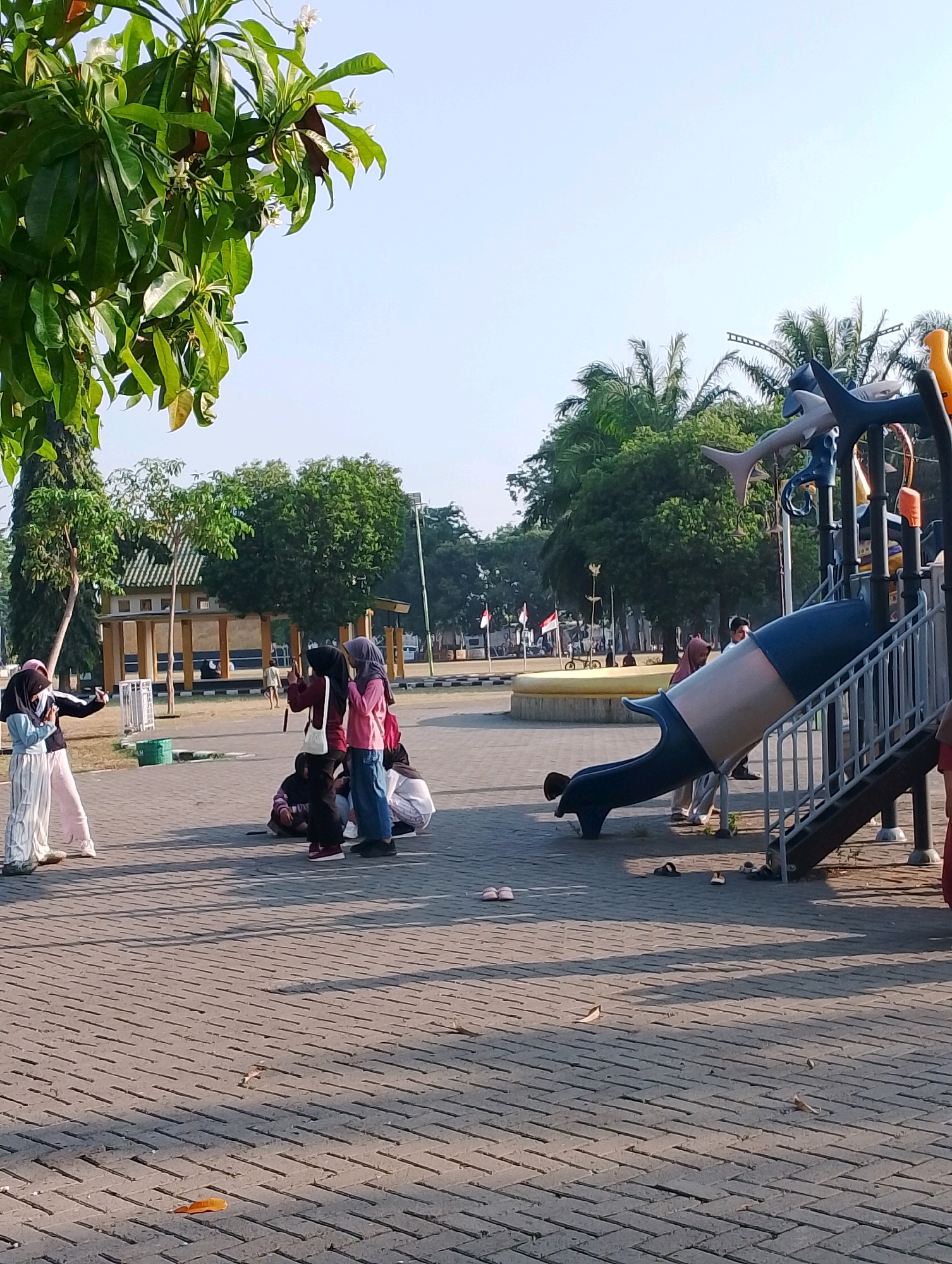 The image depicts an outdoor playground located in Probolinggo, Indonesia, as suggested by the presence of Indonesian flags and the general environment which is consistent with many public spaces in the city. The scene is set in the daytime with clear sky. In the foreground, a paved ground made of interlocking bricks forms the surface of the playground. To the left, a group of four young females are gathered. One, in the foreground and facing the viewer, is wearing a light blue hijab and striped pants, and is holding a phone as if taking a photo. Next to her, another female in a dark hijab and black jacket is also holding a phone. Behind them, two other females in hijabs are sitting on the ground.
In the mid-ground, a playground structure is visible. It features a blue and white slide, a set of stairs, and colorful decorations including shark-like figures. Several individuals are around the playground, with one person in a brown hijab and white attire standing near the slide, and another person in the background near a yellow circular platform.
The background reveals lush green trees and some architectural structures, possibly part of a park or recreational area. Indonesian flags are planted in the distance, reinforcing the location's identity. The overall impression is a lively community space, with people enjoying outdoor activities in a well-maintained public area.