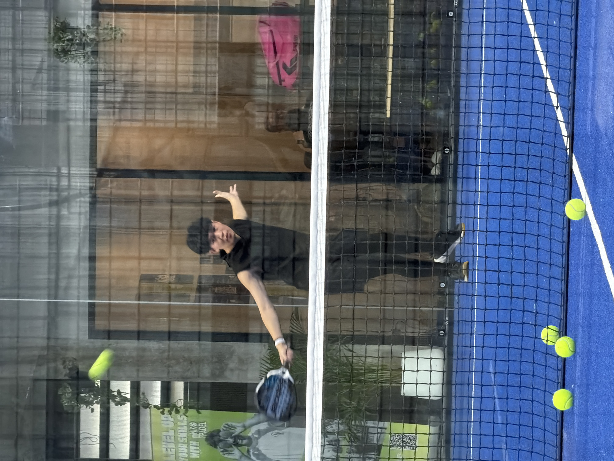 The image captures a young man playing padel on an indoor court in Ban Buak Khrok North, Thailand. The court is vibrant blue with white markings and a black net divides the playing area. The man, dressed in a black sleeveless hoodie and black athletic pants, is in mid-action, swinging a padel racket at a bright yellow ball in the air. His body is positioned to make a powerful stroke, and his gaze is focused on the ball.
The background reveals the indoor setting, with large glass panels reflecting the environment. Through the glass, we can see a potted plant with lush green leaves, a yellow poster with text that reads "LEVEL UP YOUR SKILLS WITH NIKKO PADEL," and some indistinct figures in the distance. A bright pink sports bag is also visible in the background. On the court, several yellow padel balls are scattered on the blue surface, some near the net. The lighting appears to be artificial, creating a somewhat diffused and reflective atmosphere within the court. The overall scene depicts an active sports moment within a modern indoor recreational facility.