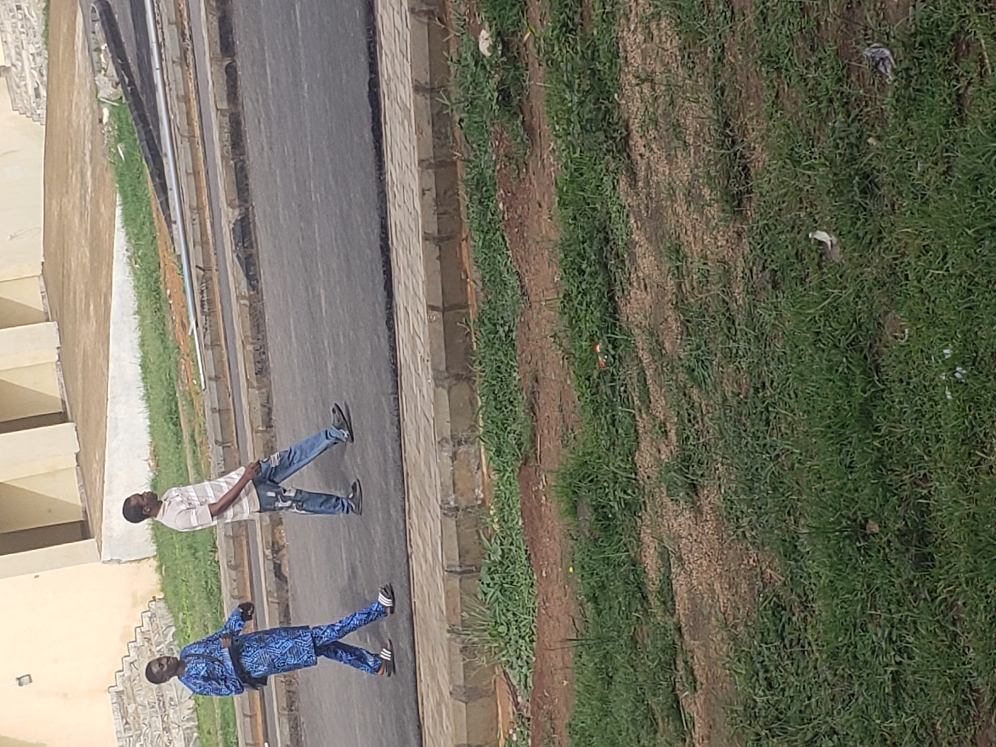 Two men are walking on an asphalt road in Gumel, Nigeria. The man on the left is wearing a blue traditional African outfit and sandals. He has a bag slung over his shoulder. The man on the right is wearing a striped t-shirt and ripped jeans, also wearing sandals. They are walking away from the camera. The road is bordered by a concrete curb and grassy areas. In the background, there is a building with a light-colored facade and a staircase.
