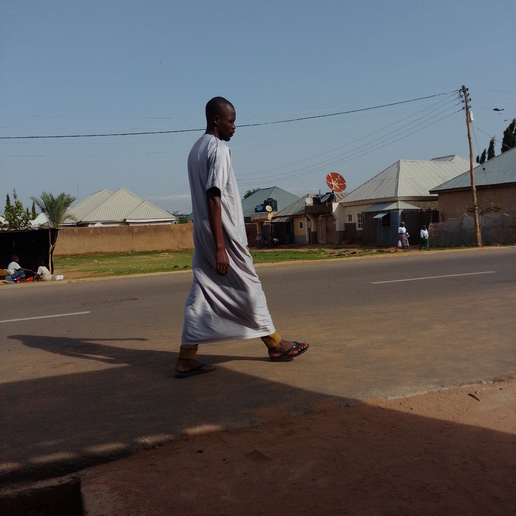 In Gombe, Nigeria, a man wearing a long white garment and brown sandals walks on an asphalt road. The man is facing right, and his shadow stretches across the road. In the background, there are houses with metal roofs, a satellite dish, and trees. The sky is clear and blue. To the left of the road, two people are sitting under a shaded structure.