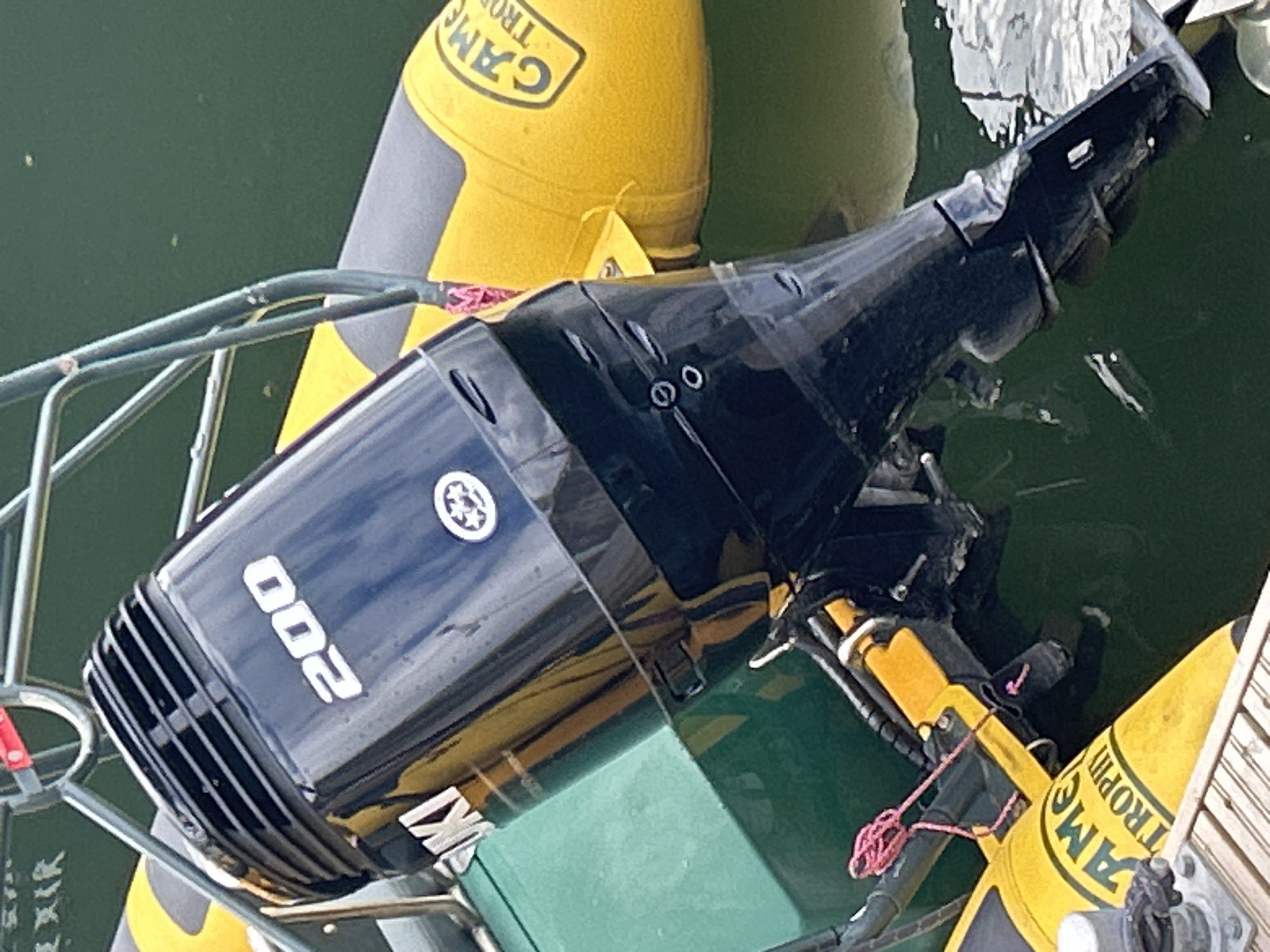 The image shows the back portion of a black Suzuki outboard motor with the number "200" prominently displayed in white lettering. The motor is mounted on the back of a green boat, which is partially submerged in dark green water. To the right of the motor, a yellow inflatable tube is visible, with the text "CAMEL TROPHY" in black letters. The overall scene suggests a marine environment, possibly a harbor or a dock. There are no people visible in the image, and no discernible actions or interactions are taking place. The image provides no specific information to locate the scene in Tallinn, Estonia.