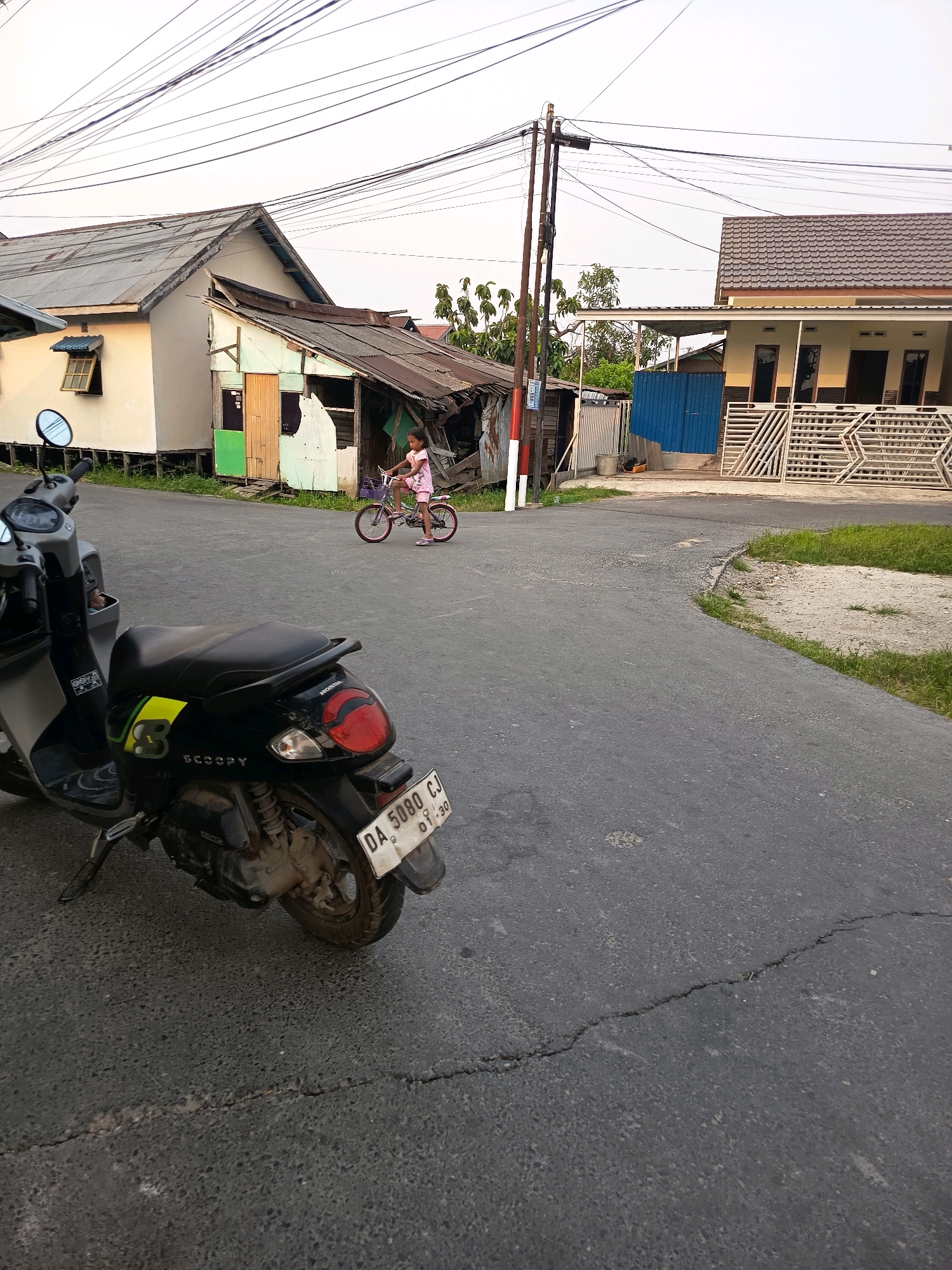 A young girl in a pink dress rides a bicycle on a paved road in Banjarmasin, Indonesia. She is located near a collection of simple houses and a modern house with a metal fence. A black scooter is parked on the left side of the road in the foreground. The scene appears to be in a residential area with overhead electrical wires visible. The sky is overcast, and the overall lighting suggests it is late afternoon or early morning. The road has some cracks and uneven surfaces.