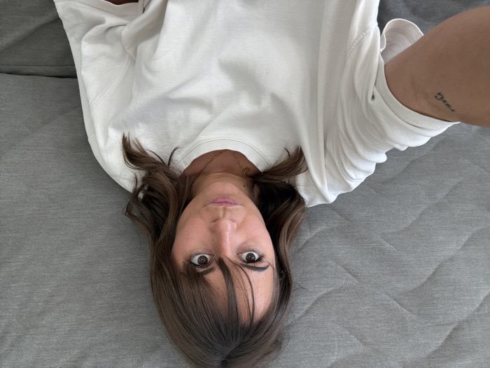 A woman lies upside down on a gray quilted surface. She has brown hair with bangs and wide, expressive eyes. She wears a white long-sleeved shirt and a delicate gold necklace. Her left arm is visible, showing a tattoo on her upper arm. The camera angle is directly above her, capturing a selfie-style perspective.
