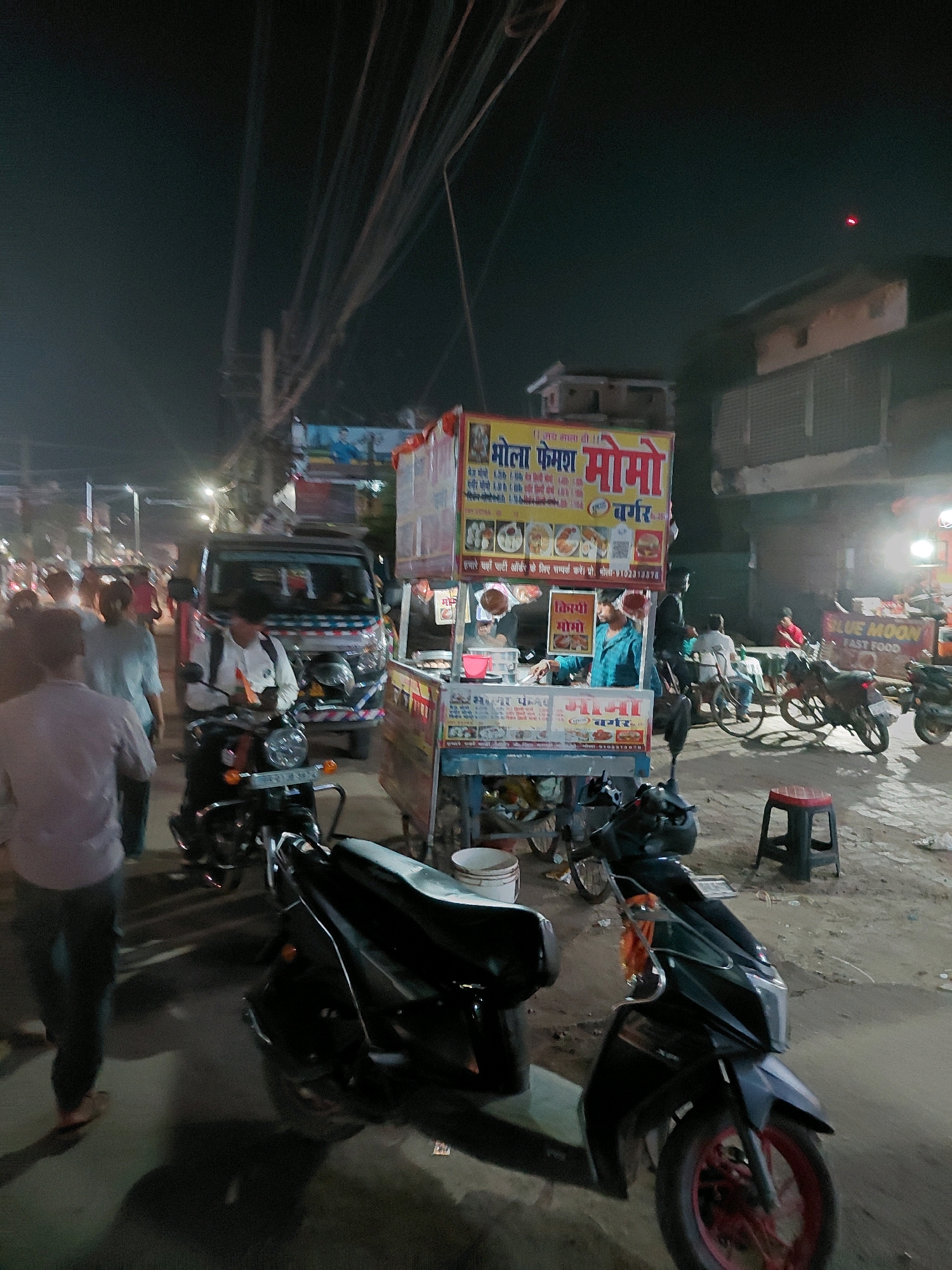 The image depicts a street scene at night in Patna, India, with a focus on a busy food stall. A prominent food cart, with "Momo" and "Burger" written in Hindi, is illuminated by lights. Several people are present, including vendors and customers, with some individuals standing and others on motorcycles. One vendor, wearing a blue shirt, is actively preparing food behind the cart. Motorcycles are parked on the street, and there is a general atmosphere of a bustling market or roadside eatery. The background shows buildings and overhead electrical wires, typical of an urban Indian setting.