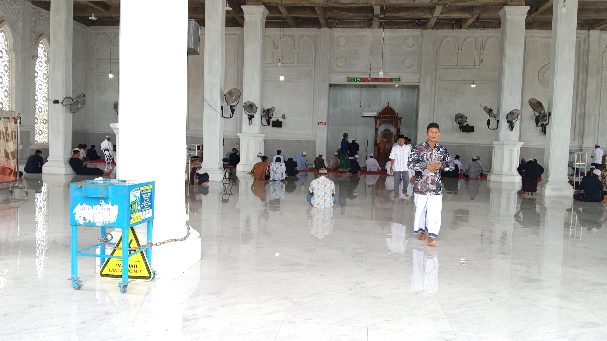 This is a wide-angle interior shot of a mosque in Bireuen, Indonesia, with a polished white marble floor reflecting the sunlight. The mosque features tall, white columns with ornate arched windows and a wooden ceiling. Multiple men are scattered across the floor, some sitting in prayer, others standing or walking. In the foreground, a blue metal cart with wheels and a yellow hazard sign is chained to a pillar. A man in a patterned shirt and white pants stands in the middle ground, facing the camera. The environment is well-lit, with natural light streaming in from the windows and ambient interior lighting.