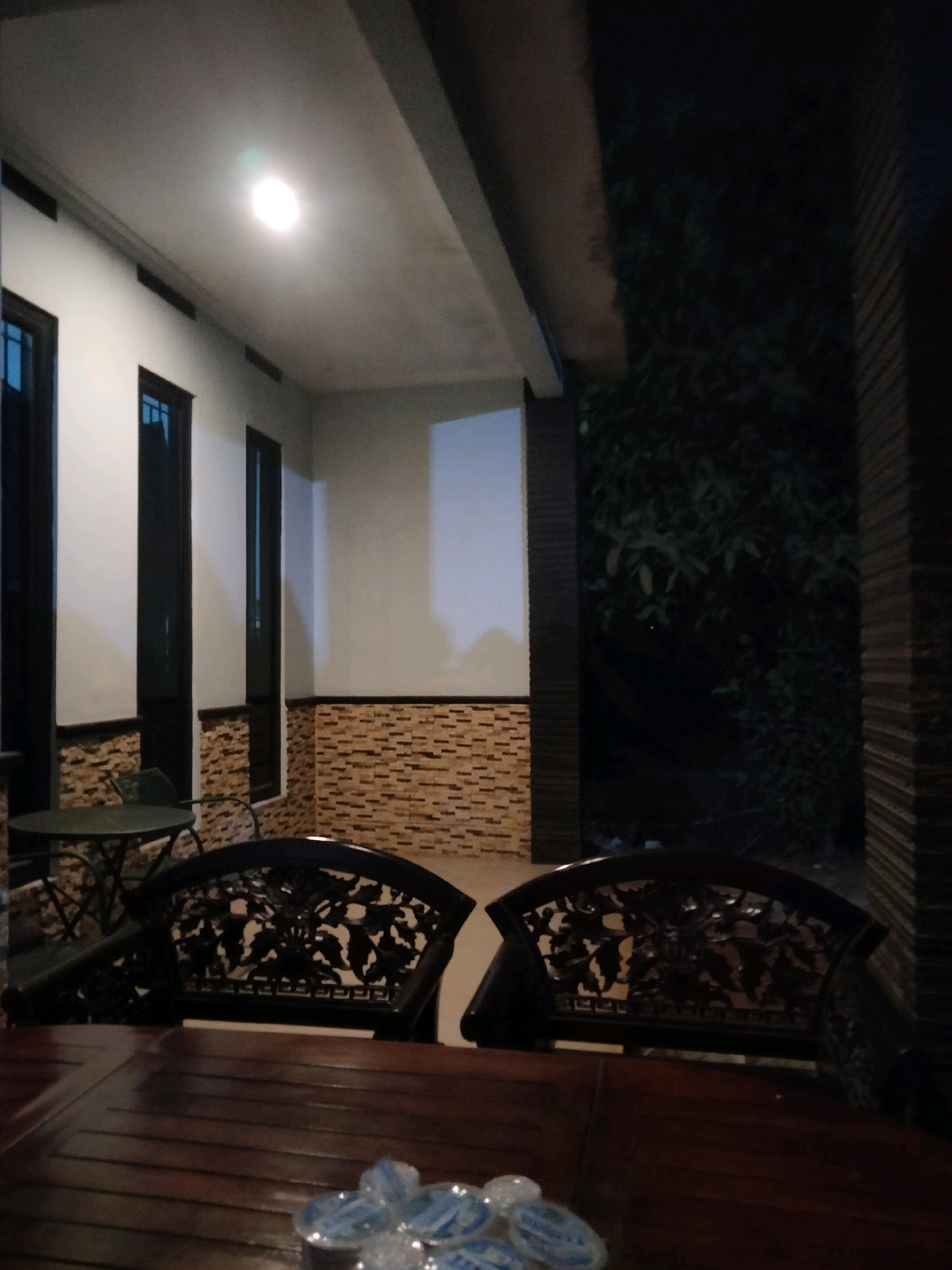 The image depicts an outdoor seating area at night, likely a cafe or restaurant, in Suka Sari, Indonesia. Two ornate dark wooden chairs are positioned in the foreground, facing a wooden table. On the table, a cluster of small, clear plastic cups containing a blue liquid is visible. In the background, a building with white walls and dark-framed windows is illuminated by an overhead light. A textured brick-like wall runs along the base of the building. To the right of the building, dense dark foliage can be seen, suggesting the presence of trees or a garden. The overall atmosphere is dim and tranquil, typical of an evening setting.