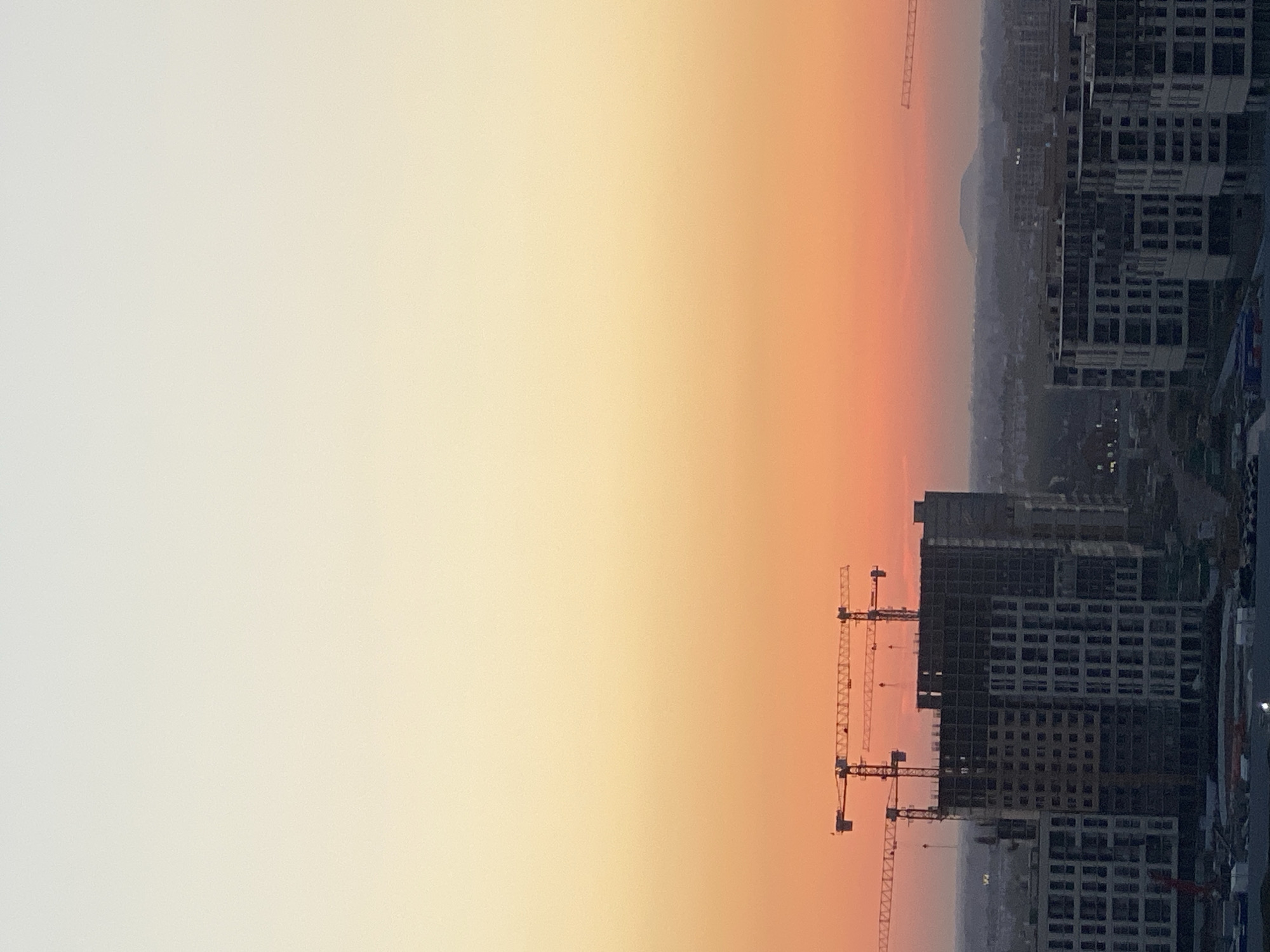An aerial photograph captures a cityscape at dusk. The sky is a gradient of soft yellows and oranges, transitioning into a pale peach hue near the horizon, suggesting the last moments of daylight. In the foreground, construction cranes are silhouetted against the vibrant sky, hovering over several partially built buildings. These structures appear to be modern residential or commercial complexes, with visible scaffolding and exposed concrete. The buildings are clustered together, indicating a developing urban area. In the background, a hazy silhouette of a mountain or hill is visible, partially obscured by the atmospheric conditions. The overall scene depicts a city undergoing expansion and development, bathed in the warm glow of a sunset. The location is Novotitarovskaya, Russia.