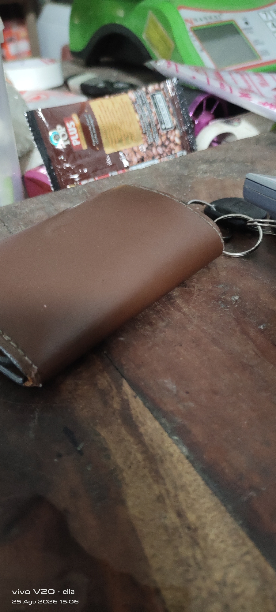 The image shows a brown leather coin purse and a set of keys on a wooden surface. In the background, a packet of "TOP PLUS" coffee is visible, along with other items on a desk or table. The timestamp at the bottom left corner indicates the image was taken on August 25, 2026, at 15:06, with the device "vivo V20". The location of Simo, Indonesia, is also mentioned. The scene appears to be an indoor setting, possibly a home or office. There are no people visible in the image.