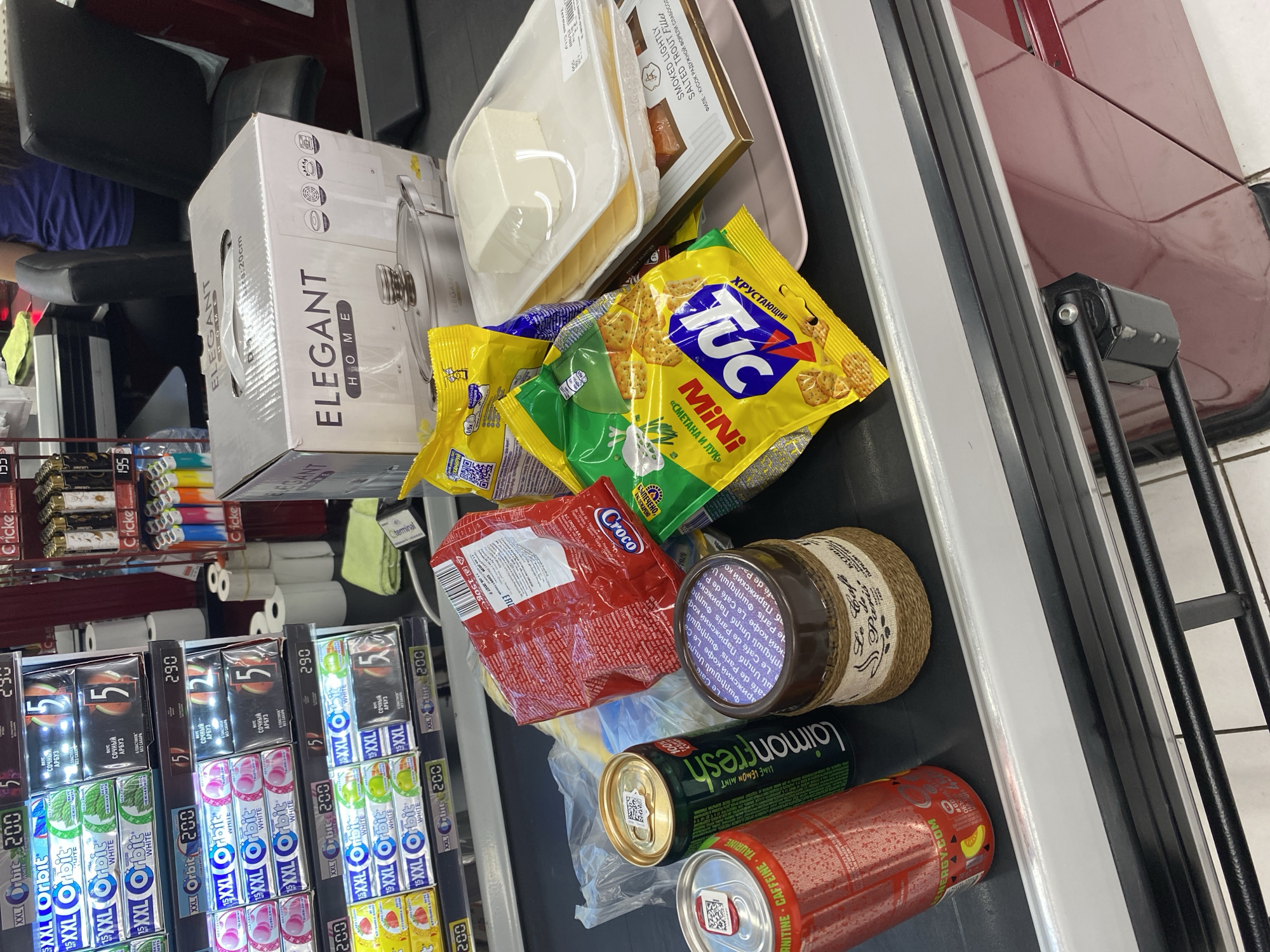 The image shows a checkout counter at a supermarket in Yerevan, Armenia. Several grocery items are placed on the conveyor belt, including cans of energy drinks, packages of crackers and snacks, a jar of coffee, and containers of cheese and smoked trout. A cardboard box for a "Elegant Home" pot is also visible. In the background, shelves stocked with gum and other products can be seen, along with a cashier and a customer. The overall scene depicts a typical retail environment.