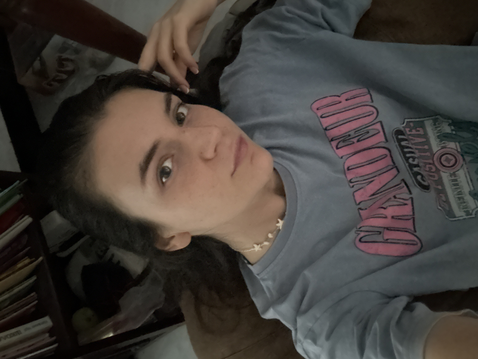 A woman is lying down, wearing a light blue t-shirt with a pink and gray graphic design. She has dark hair, fair skin with freckles, and is wearing a delicate necklace with small, white star-shaped charms. Her left hand is near her head. A bookshelf with books and other objects is visible on the left. There is no information about Mũi Né, Vietnam in this image.