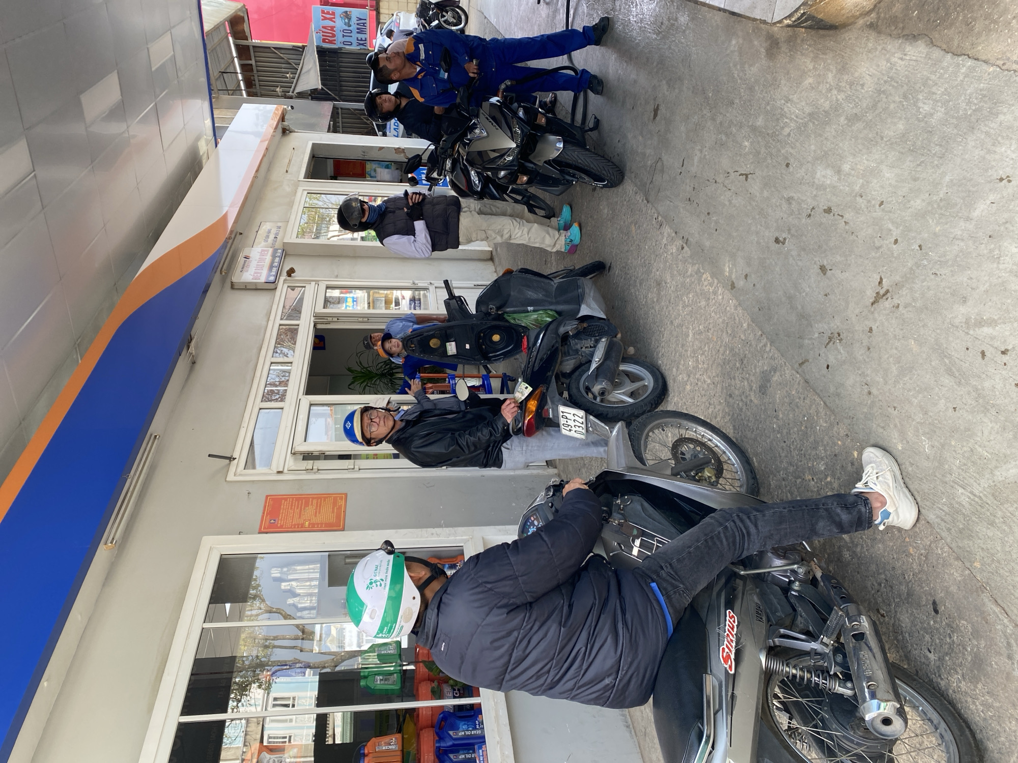 A gas station scene in Lam Vien - Da Lat, Vietnam. Four men are present, two wearing blue uniforms, presumably gas station attendants. The other two men are on motorbikes. The man closest to the camera wears a black jacket and a helmet with green and white accents; the other has a blue helmet and glasses. The motorcycle closer to the camera has a "Sirius Yamaha" sticker, and a license plate "49-P1 0322" is visible on the other one. The gas station storefront includes a sign that says "RUA XE O TO XE MAY". Bottles of oil are visible on a shelf behind the person on the motorcycle closer to the camera.