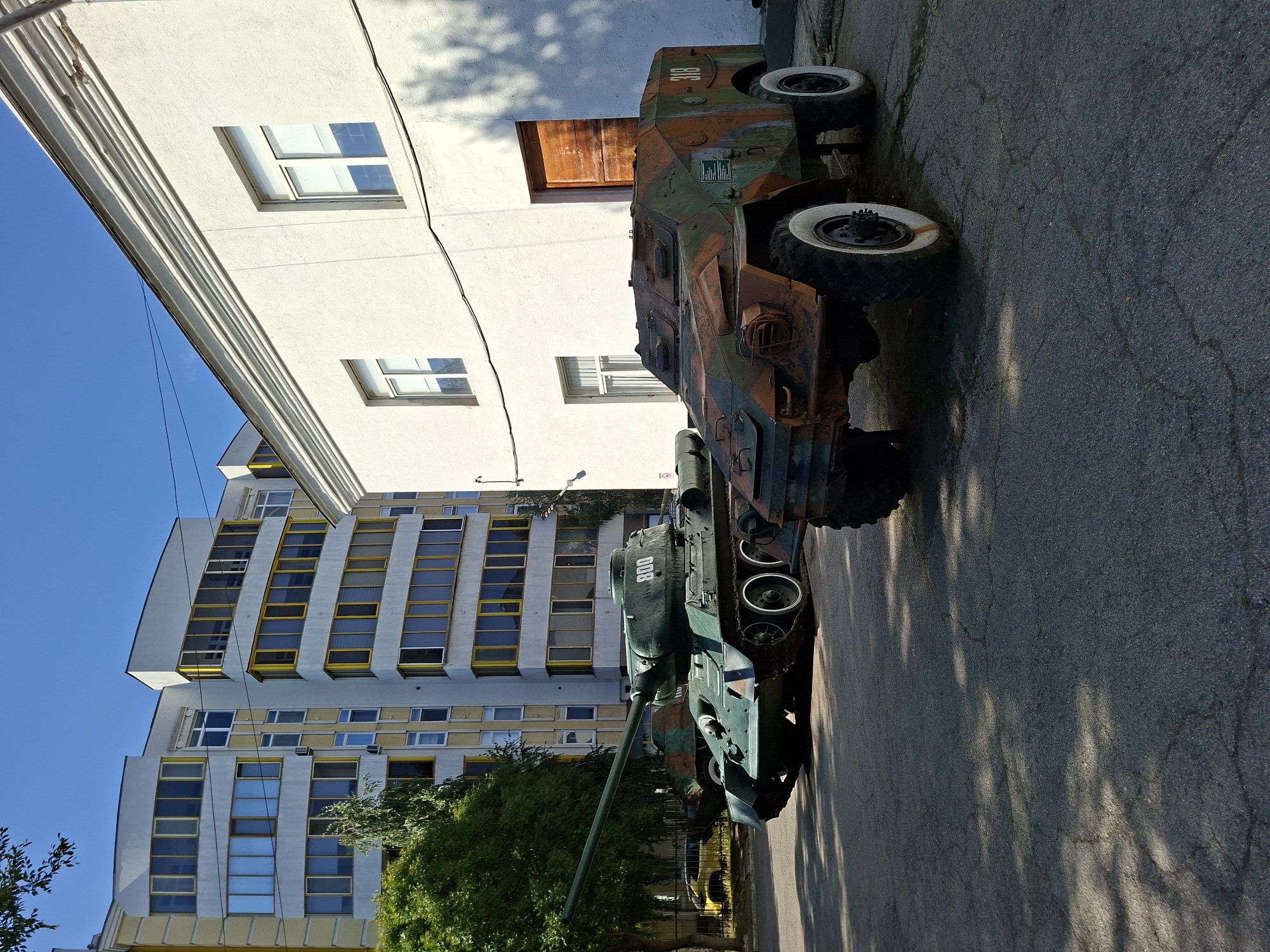 The image depicts a sunny outdoor scene in Chișinău, Moldova, featuring two military vehicles displayed on an asphalt surface in front of a white building and a modern yellow and white apartment building. In the foreground, to the right, is an armored reconnaissance vehicle, camouflaged with rust-colored, green, and brown splotches, with the number "318" visible on its side. It has four large wheels with white rims. To the left of the armored vehicle, and slightly behind it, is a green military tank with a long barrel. The tank has the number "800" painted on its turret. The apartment building in the background has a multi-story facade with numerous windows, some featuring yellow frames. The white building to the right appears to be a single-story structure with a wooden door and windows. The sky is clear and blue. Trees with green foliage are visible on the left side of the image, casting shadows on the ground. The scene appears to be a static display, possibly in a museum or memorial area.