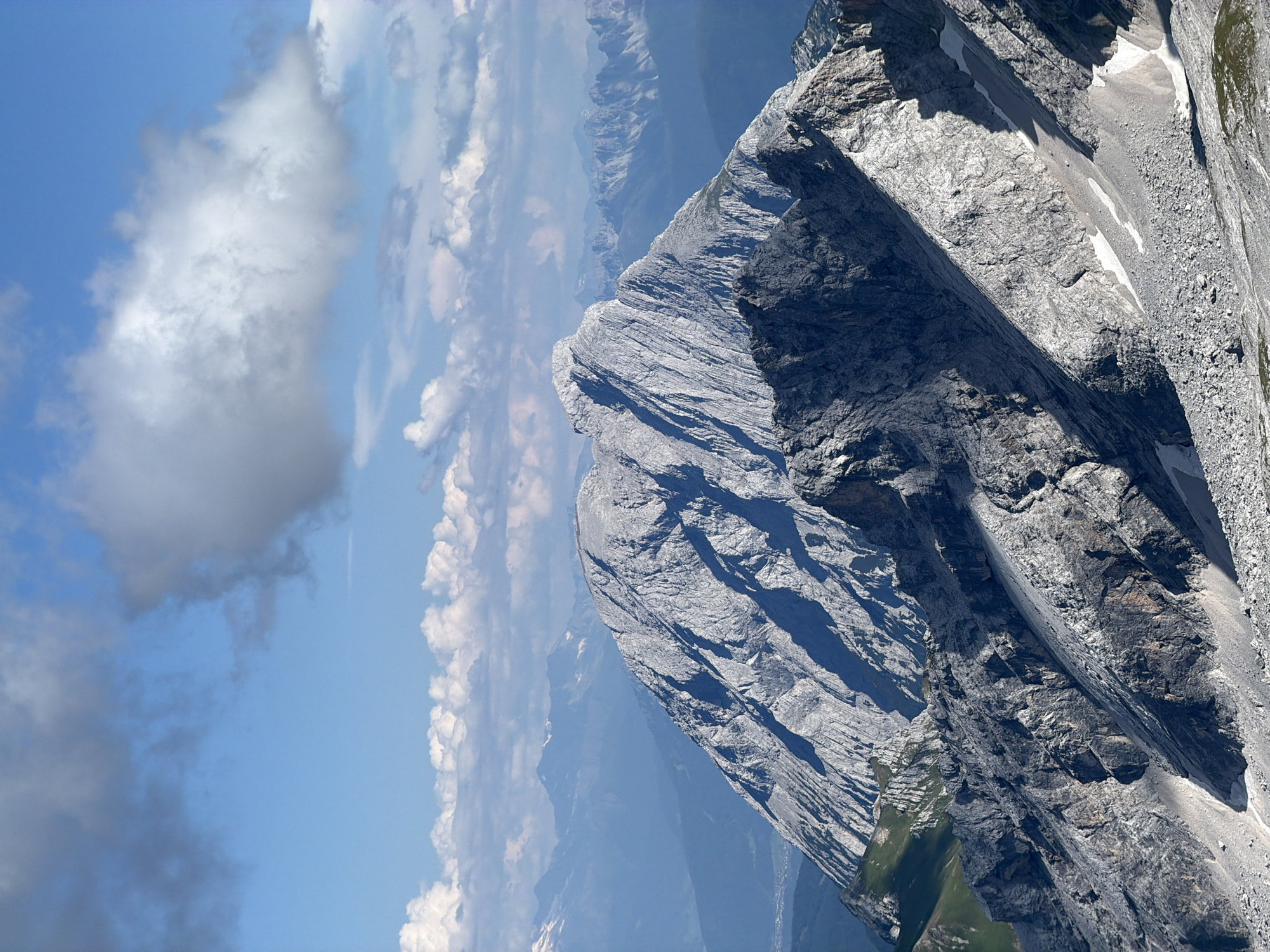 This is a vertical, eye-level, full shot of the Zugspitze mountain range, located in Garmisch-Partenkirchen, Germany. The foreground is dominated by rugged, gray, rocky slopes with patches of snow. In the midground, a large, imposing mountain with sharp ridges and deep shadows rises prominently, its summit a jagged grey mass. Further in the background, multiple layers of mountains recede into a hazy blue distance, their peaks softened by atmospheric perspective. The sky above is a vibrant blue, with a large, fluffy white cloud dominating the upper left quadrant and smaller, scattered clouds stretching across the horizon. No people or man-made objects are visible in the frame. The environment is a natural mountain landscape under a clear, partly cloudy sky.