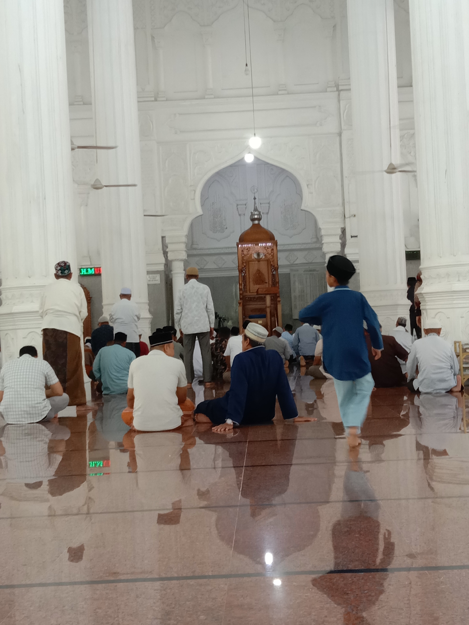 A medium shot depicts the interior of what appears to be a mosque. Several individuals are present, mostly men and one child. The men are dressed in traditional Muslim attire, including white shirts and caps. Most are seated on the glossy, reflective floor, which appears to be made of a reddish-brown stone. In the foreground, a young boy wearing a blue shirt and light blue pants is walking towards the left, his shadow reflecting on the floor.
In the background, there is an elaborately decorated white wall with arched doorways and pillars, characteristic of mosque architecture. A wooden structure, possibly a minbar, stands in the center of the background. The interior is illuminated by artificial light from hanging lamps and ceiling fixtures.
The location is indicated to be Lhokseumawe, Indonesia. The activities suggest a religious gathering or prayer session.