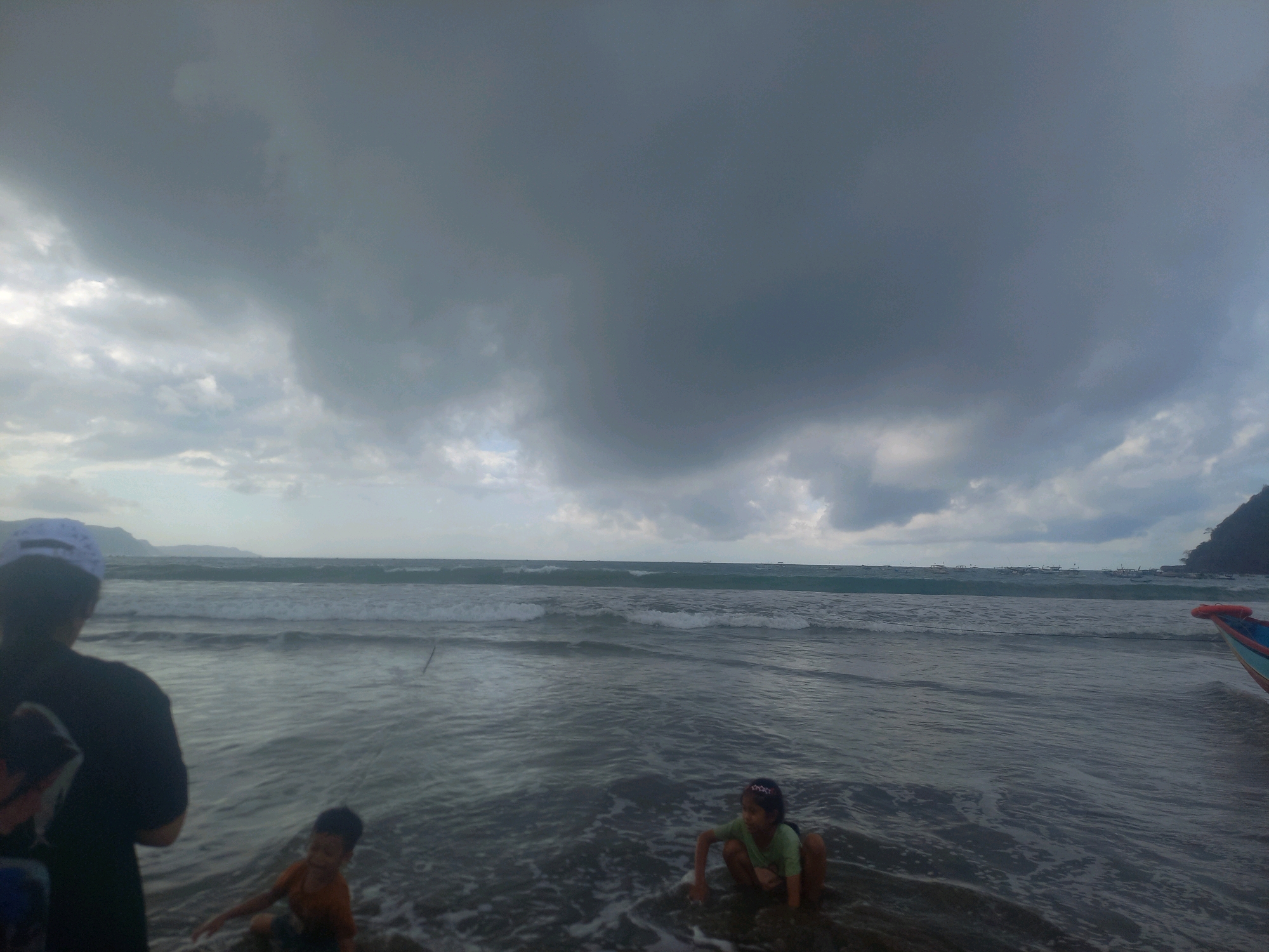 An eye-level, wide shot captures a cloudy day at a beach in Unknown, Indonesia. The sky is dominated by dark, imposing storm clouds that obscure much of the horizon, with hints of lighter clouds in the distance. A choppy sea stretches out, with visible waves breaking on the shore. In the foreground, the murky water laps at a dark sandy beach.
On the left, a person wearing a dark shirt and a white cap stands facing away from the camera, looking towards the sea. To their right and slightly in front, a young child in an orange shirt is visible, partially submerged in the water. Further to the right, another young child in a green shirt is crouched down in the shallow water, looking towards the left. In the far right of the frame, the colorful bow of a small boat, possibly a fishing boat, with a life preserver, is visible. In the distance, several small boats are scattered across the sea, and a dark, mountainous shoreline is faintly visible under the stormy sky. The overall atmosphere is moody and somewhat ominous due to the heavy cloud cover.